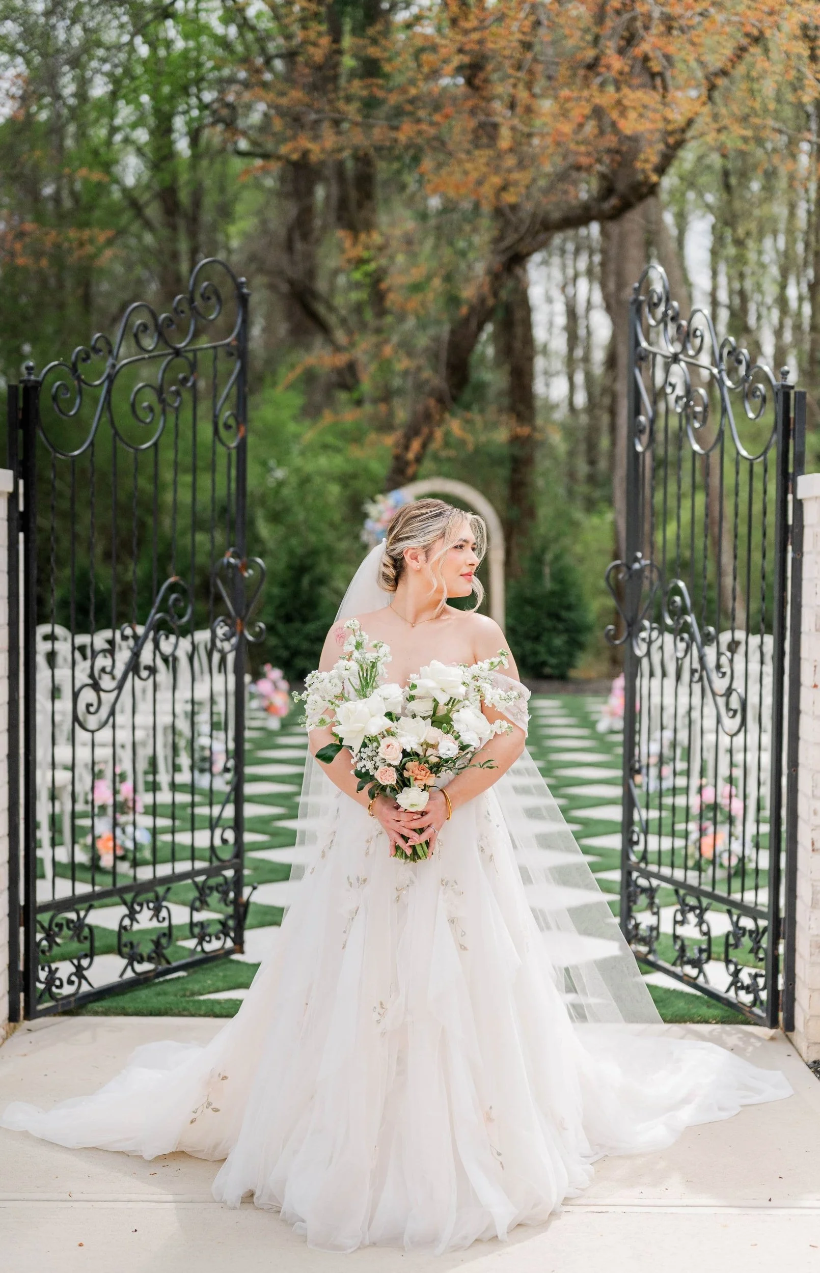 A bride in a white wedding gown holding a bouquet of white and peach flowers standing at an open wrought iron gate in a garden with trees in the background.