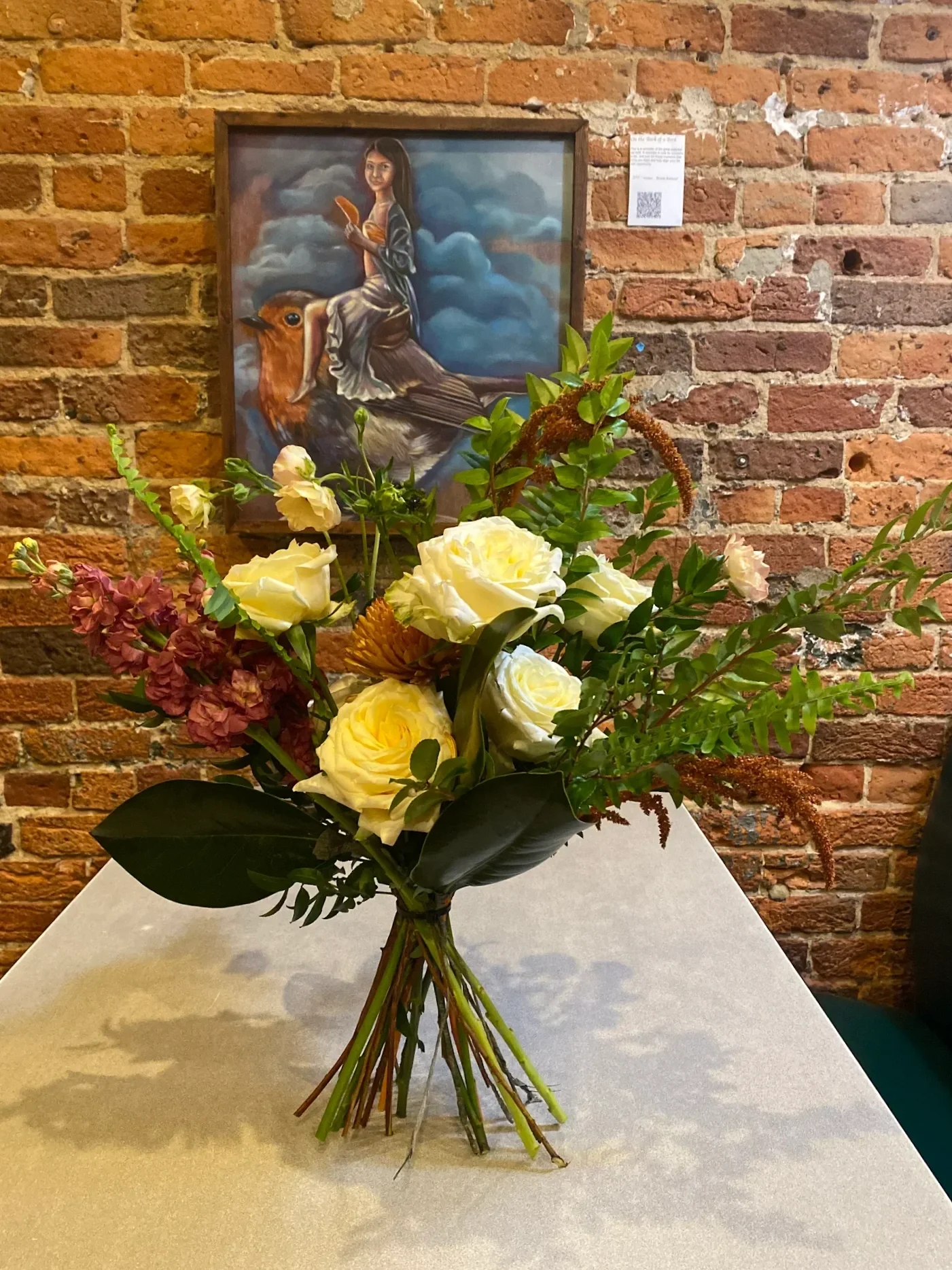 A bouquet of assorted flowers including yellow roses, pinkish hydrangeas, and green foliage on a table with a brick wall in the background. A framed art piece hangs on the brick wall depicting a woman with dark hair in a gray dress sitting on a large bird with a background of clouds.