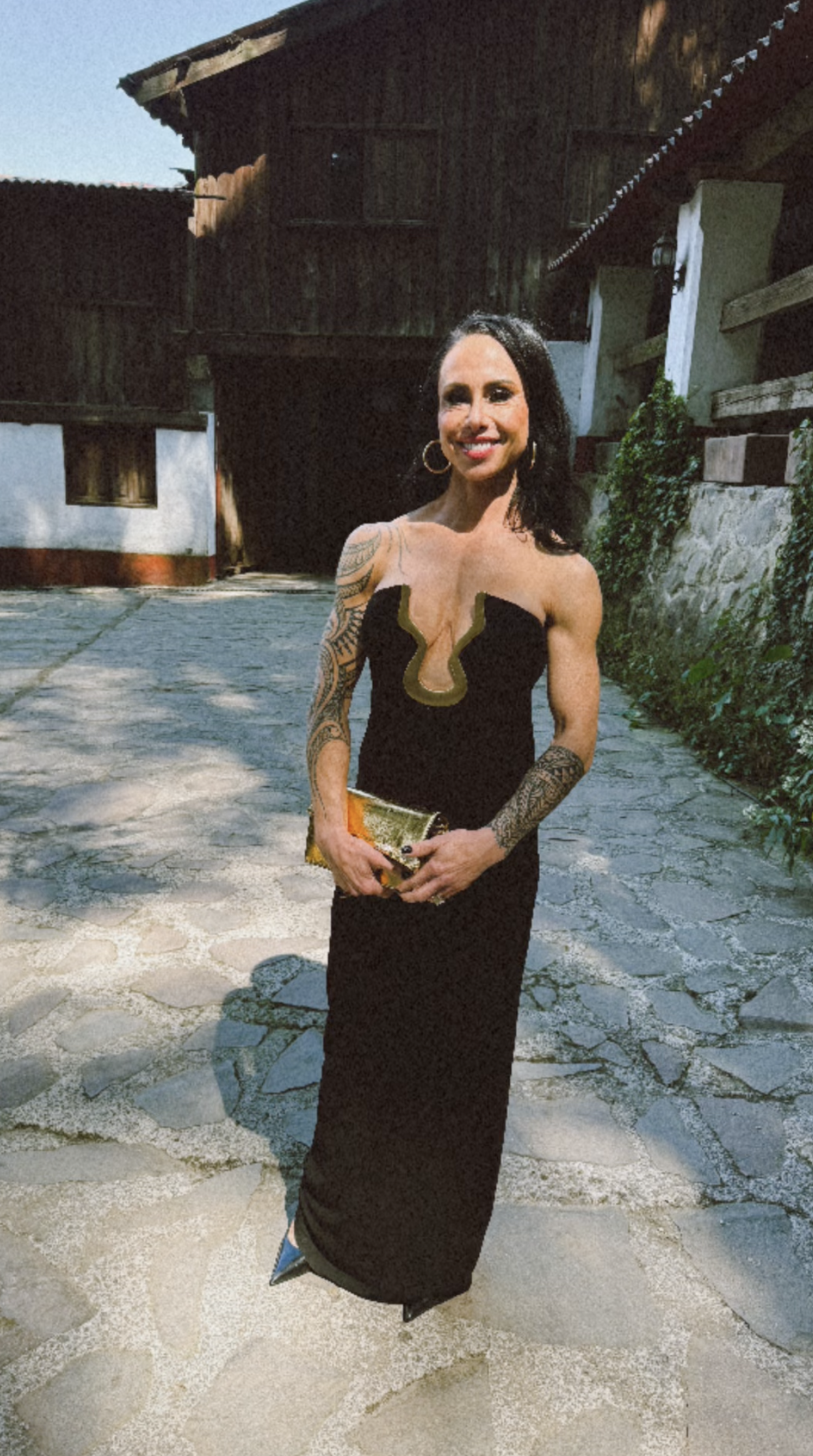 Dr. Gabrielle Lyon in a long black dress with gold trim and tattoos on her arms, standing outdoors on a stone-paved area with a wooden building in the background, smiling at the camera. Styled By: Annebet Duvall