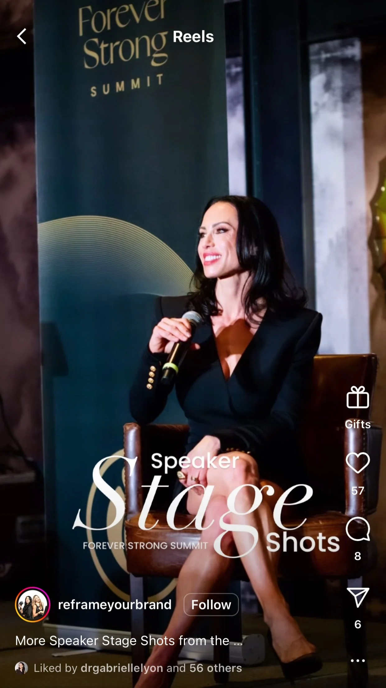 Dr. Gabrielle Lyon with black hair and a black blazer-style dress is sitting on a chair holding a microphone during a presentation at the Forever Strong Summit. There is a large blue banner beside her with the event name and logo. Styled By: Annebet