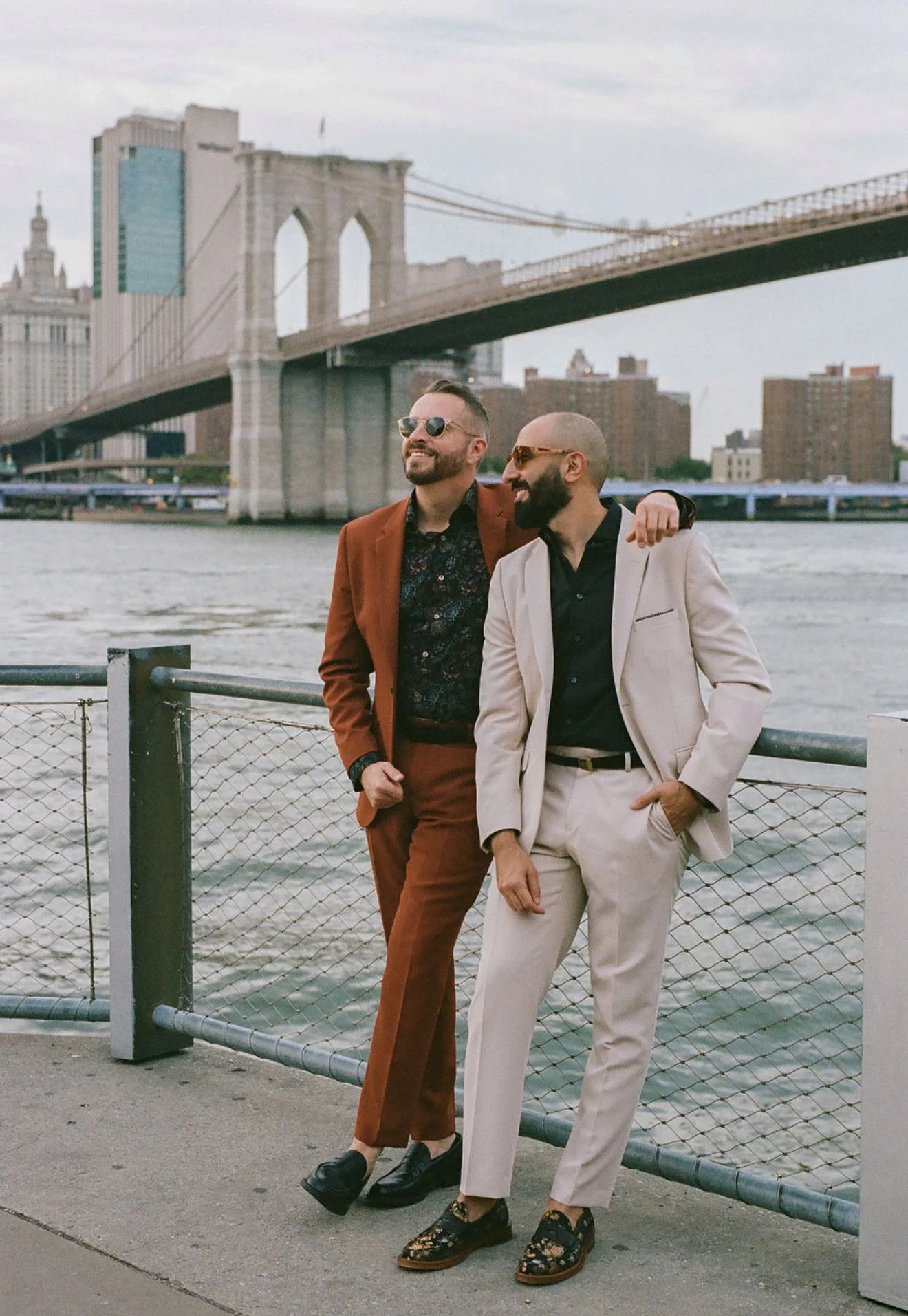 The Brownstone Boys in suits standing by a river with the Brooklyn Bridge in the background, one with an arm around the other's shoulder, both smiling and wearing sunglasses. Styled By: Annebet Duvall