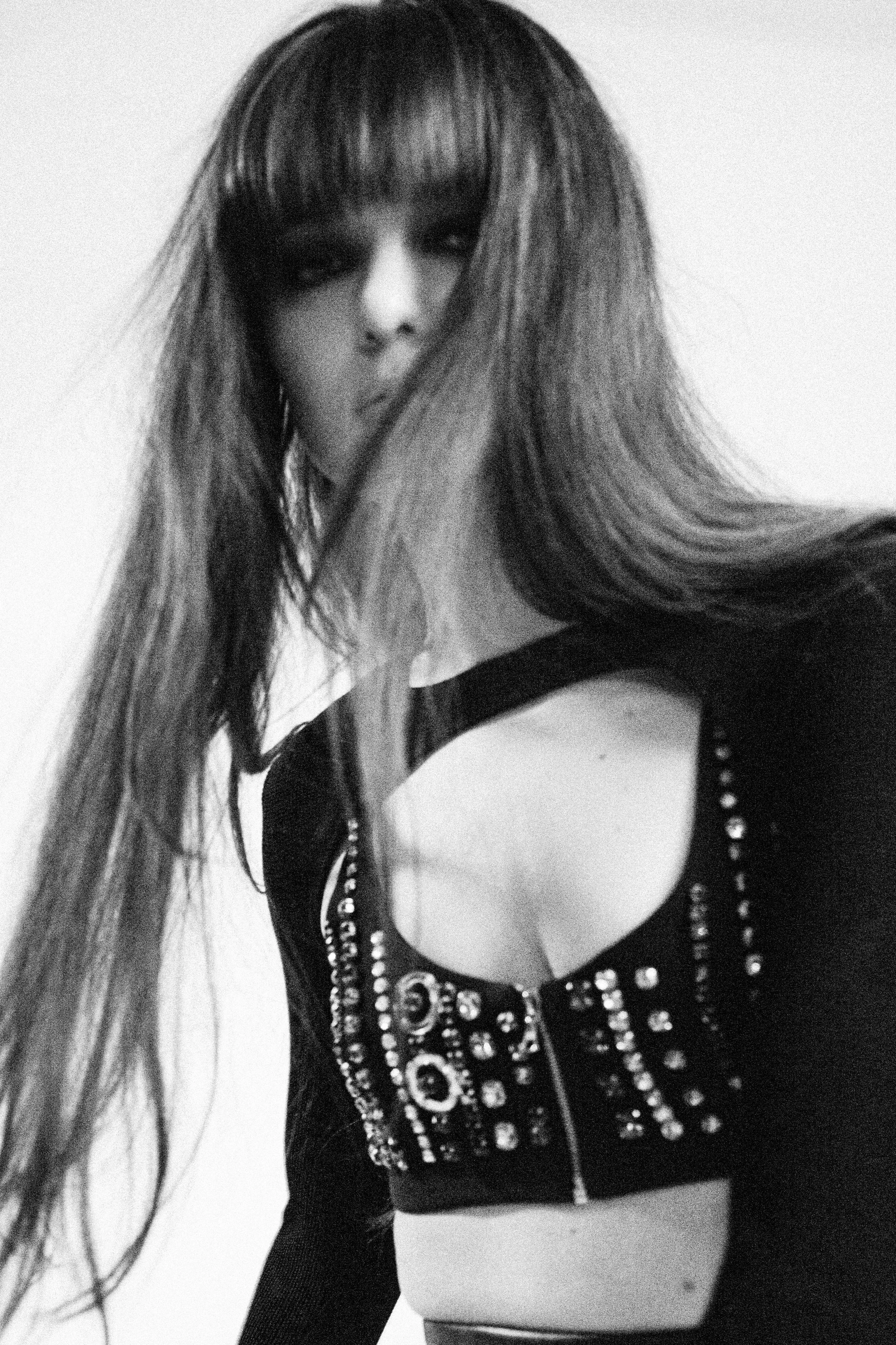 DJ Callie Reiff A with long hair and bangs wearing a black top decorated with rhinestones, posing in front of a plain white background. Styled By: Annebet Duvall