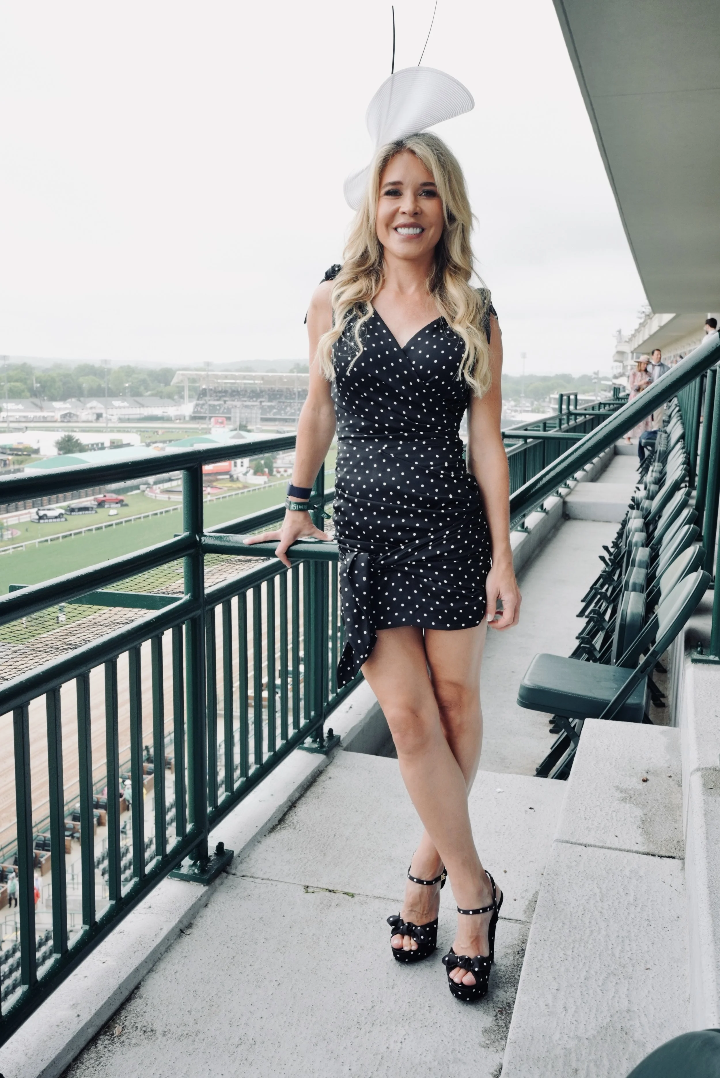 Jenn Drummond in a black polka dot Dolce and Gabbana dress and high heels stands on a balcony at a racetrack, smiling at the camera. She wears a large white fascinator hat and has blonde hair. Styled By: Annebet Duvall