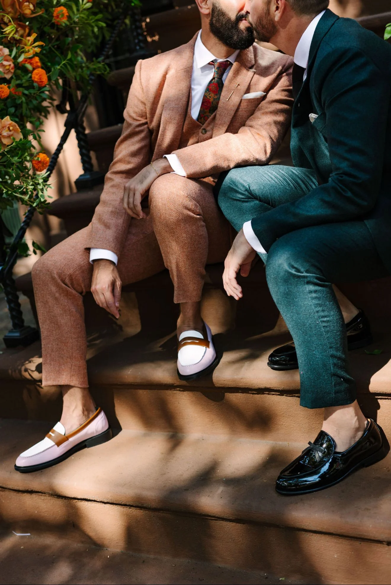 The Brownstone Boys dressed in suits sitting on a bench, leaning in as if whispering or sharing a kiss, with one man wearing pink loafers and the other black leather loafers. There are plants and flowers nearby. Styled By: Annebet Duvall