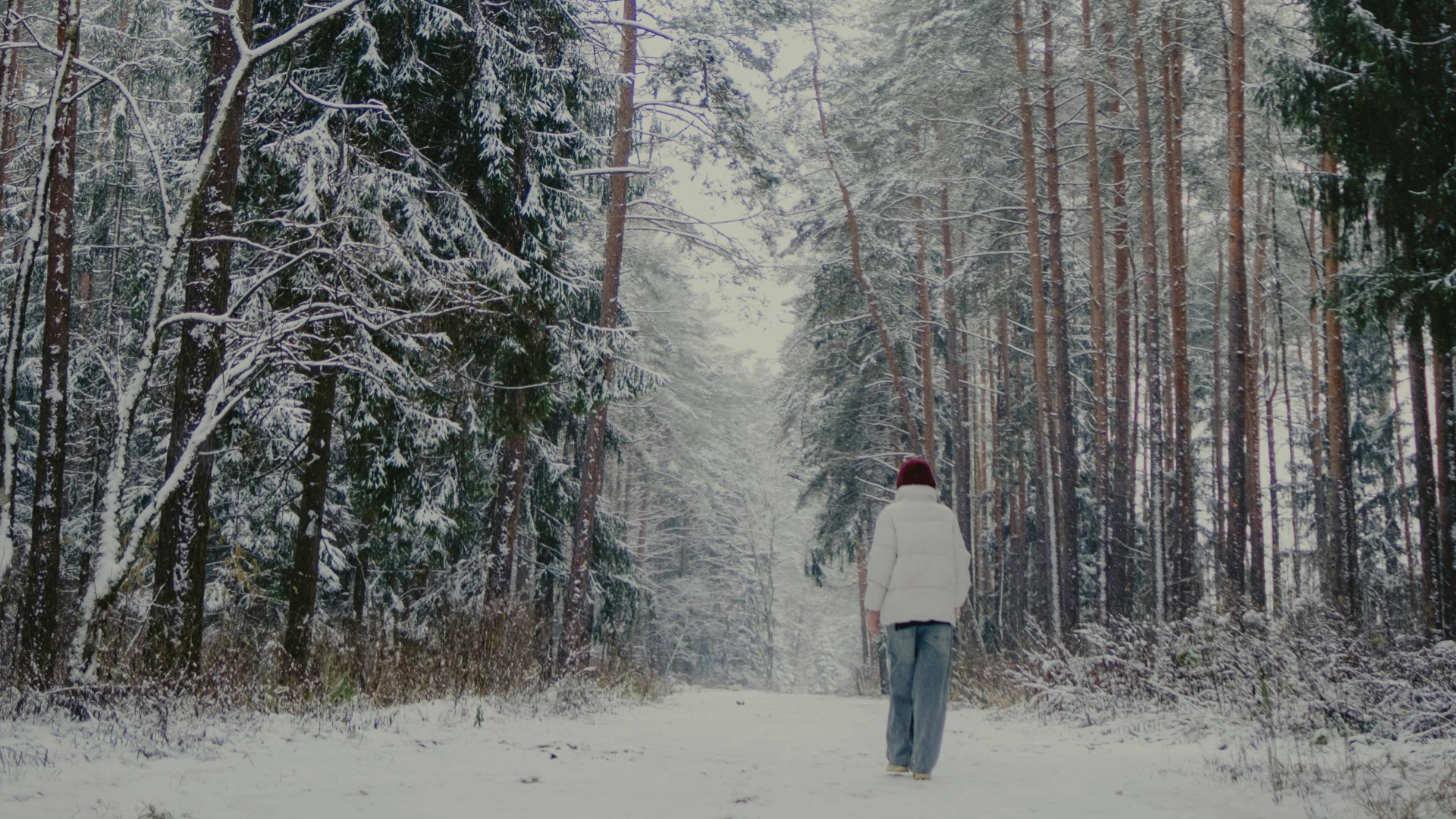 A person wearing a white winter puffer jacket, red touque, and jeans walks through a snowy forest