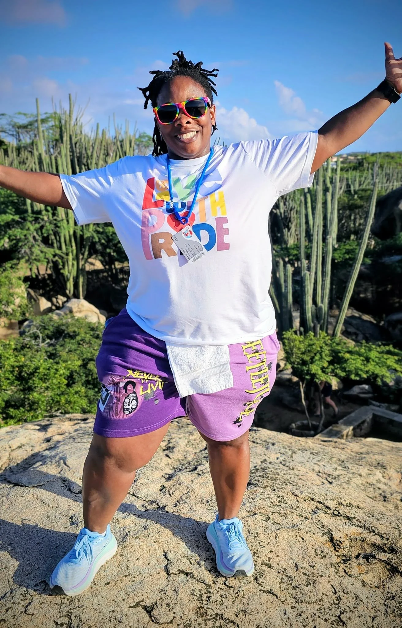 A woman smiling wearing sunglasses, a colorful tie-dye T-shirt, denim shorts, and checkered slip-on shoes, standing in a parking lot with cars and trees in the background.