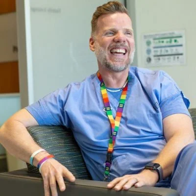 A man with short hair and a beard wearing blue scrubs and a rainbow lanyard, smiling and sitting in a hospital or medical office.