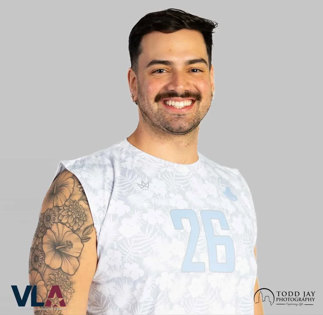 A person with red hair and glasses wearing a gray leather jacket outdoors with a river, mountains, and autumn trees in the background.