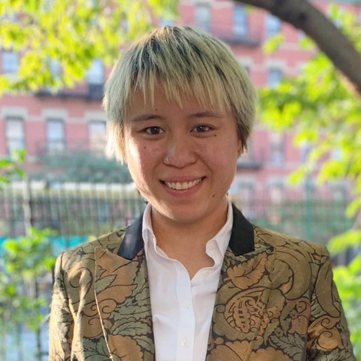 Woman with curly hair, glasses, and a black shirt with rainbow-colored text, smiling and flashing peace signs.
