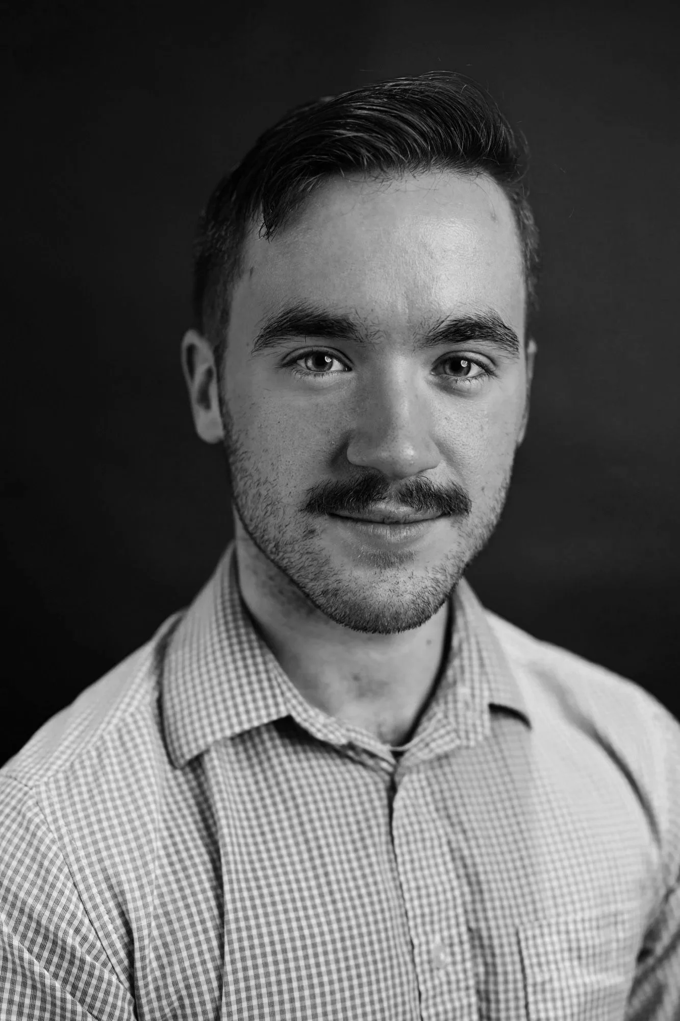 Black and white portrait of a young man with short dark hair, a mustache, and a goatee, wearing a checkered shirt.