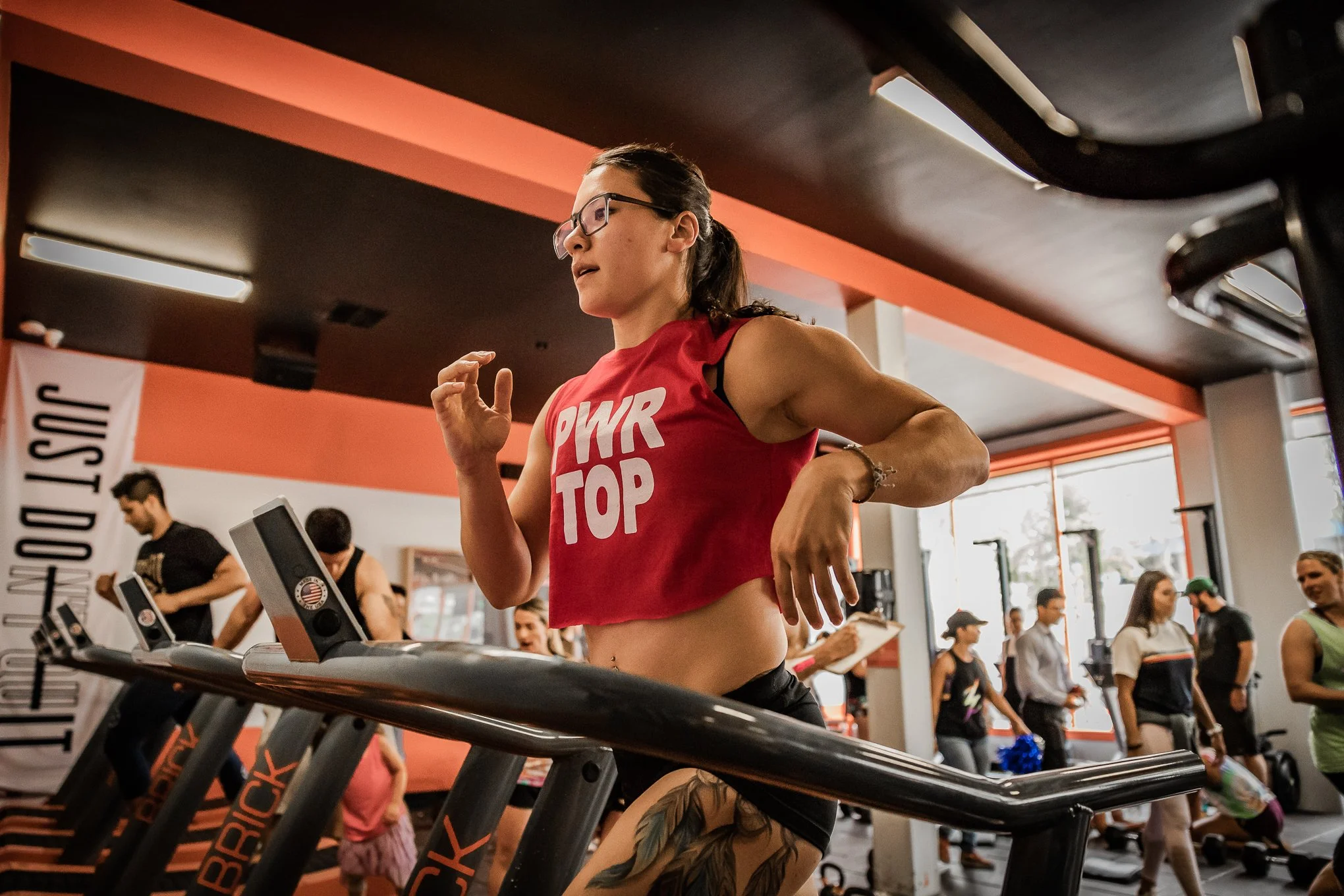 A woman with glasses and a tattoo on her thigh, wearing a red sports top that says 'PWR TOP,' is running on a treadmill at a gym, with other people exercising in the background.