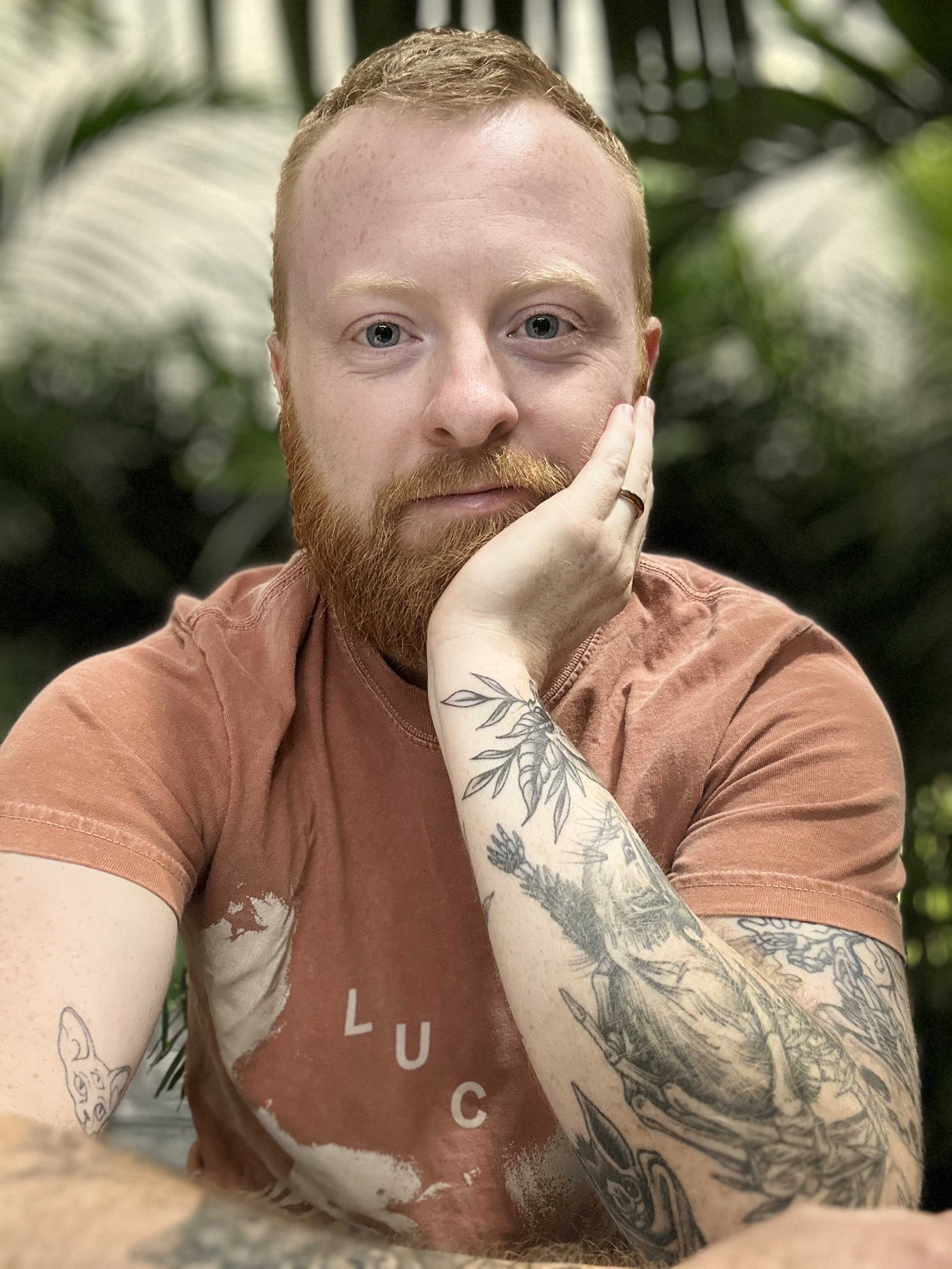 A young man with red hair and beard wearing a brown T-shirt, jewelry, and tattoos, standing against a black background.