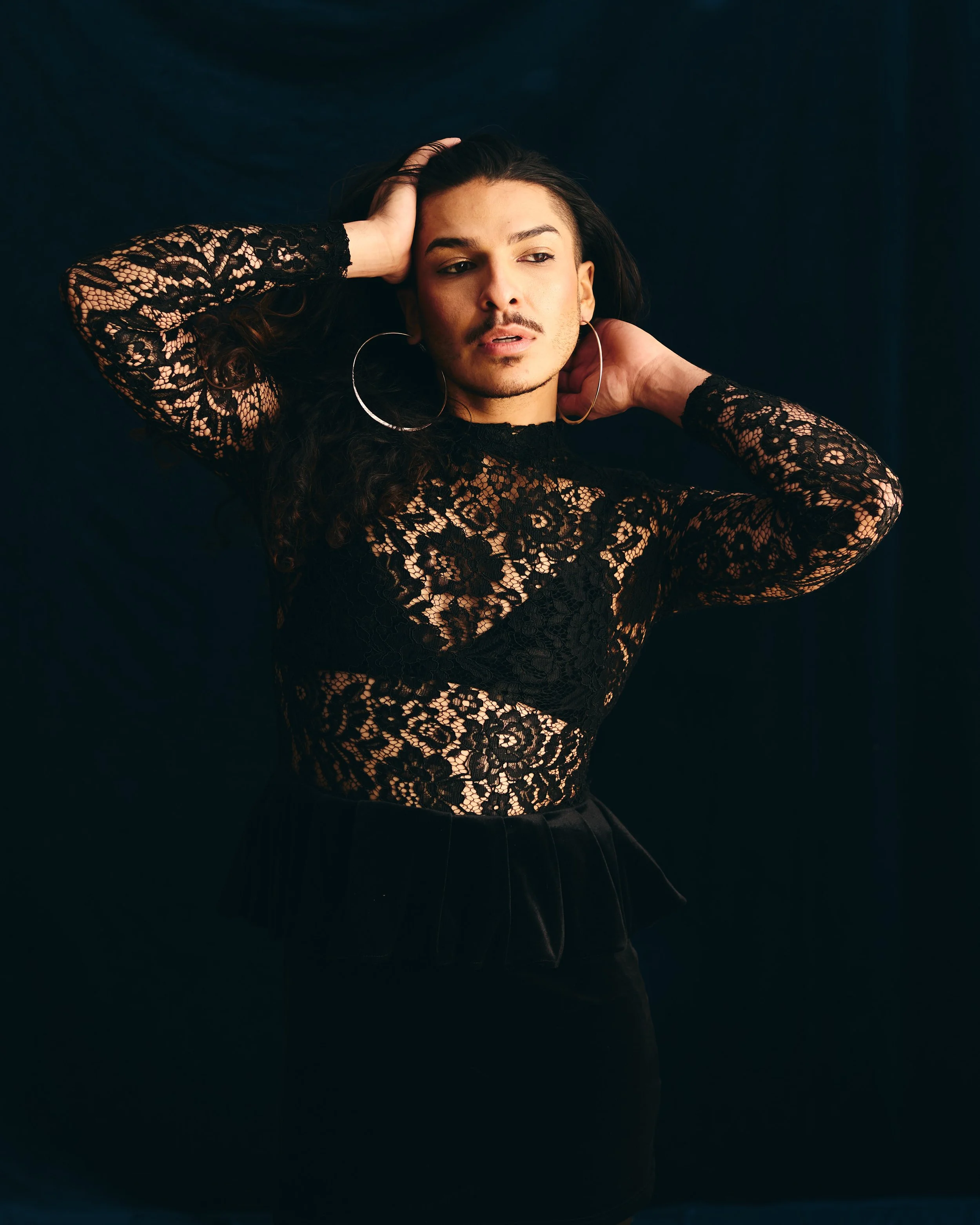 A person with shoulder-length wavy hair, wearing a black blazer and a pearl necklace, posing against a warm, reddish-brown background.
