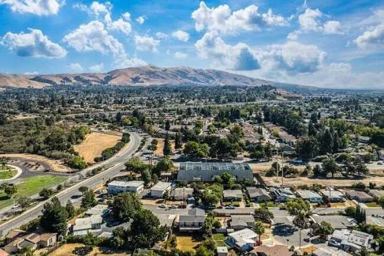 Aerial view of a suburban neighborhood with houses, roads, and greenery, with mountains in the background and a partly cloudy sky.