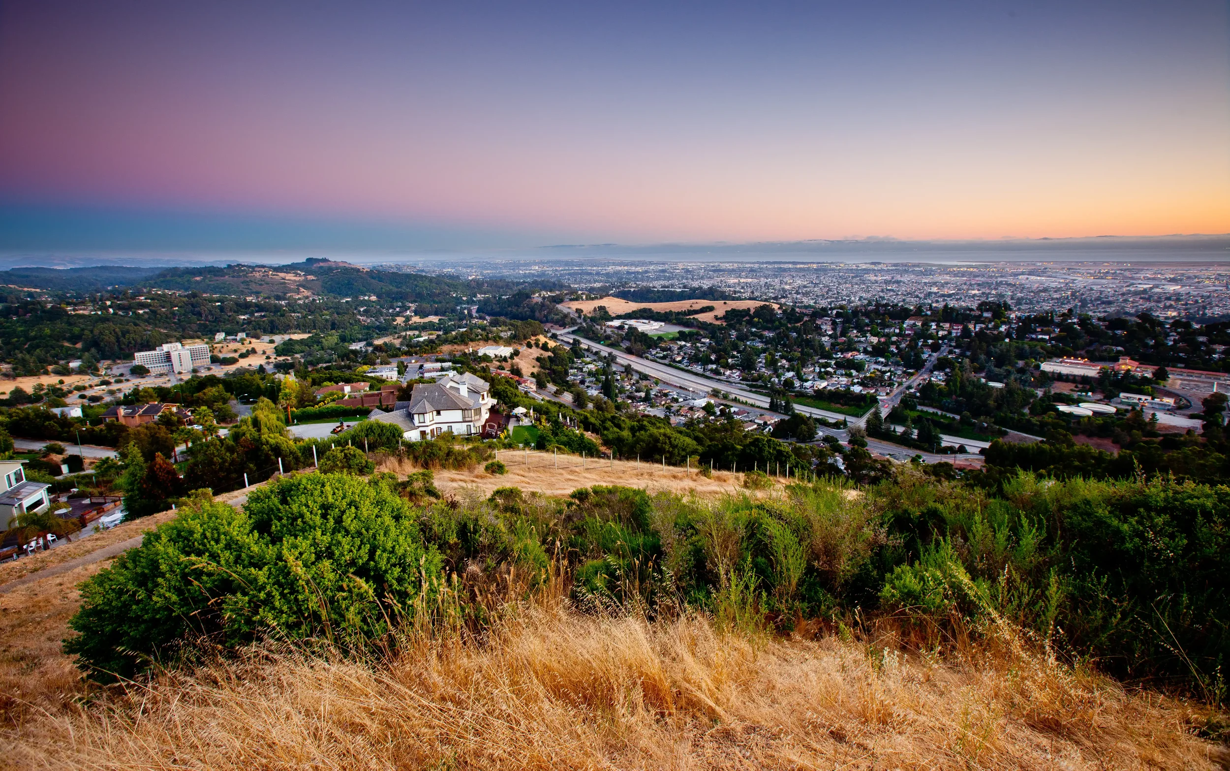 A scenic view of a city from a hillside at sunset, with houses, trees, highways, and urban landscape under a colorful sky.