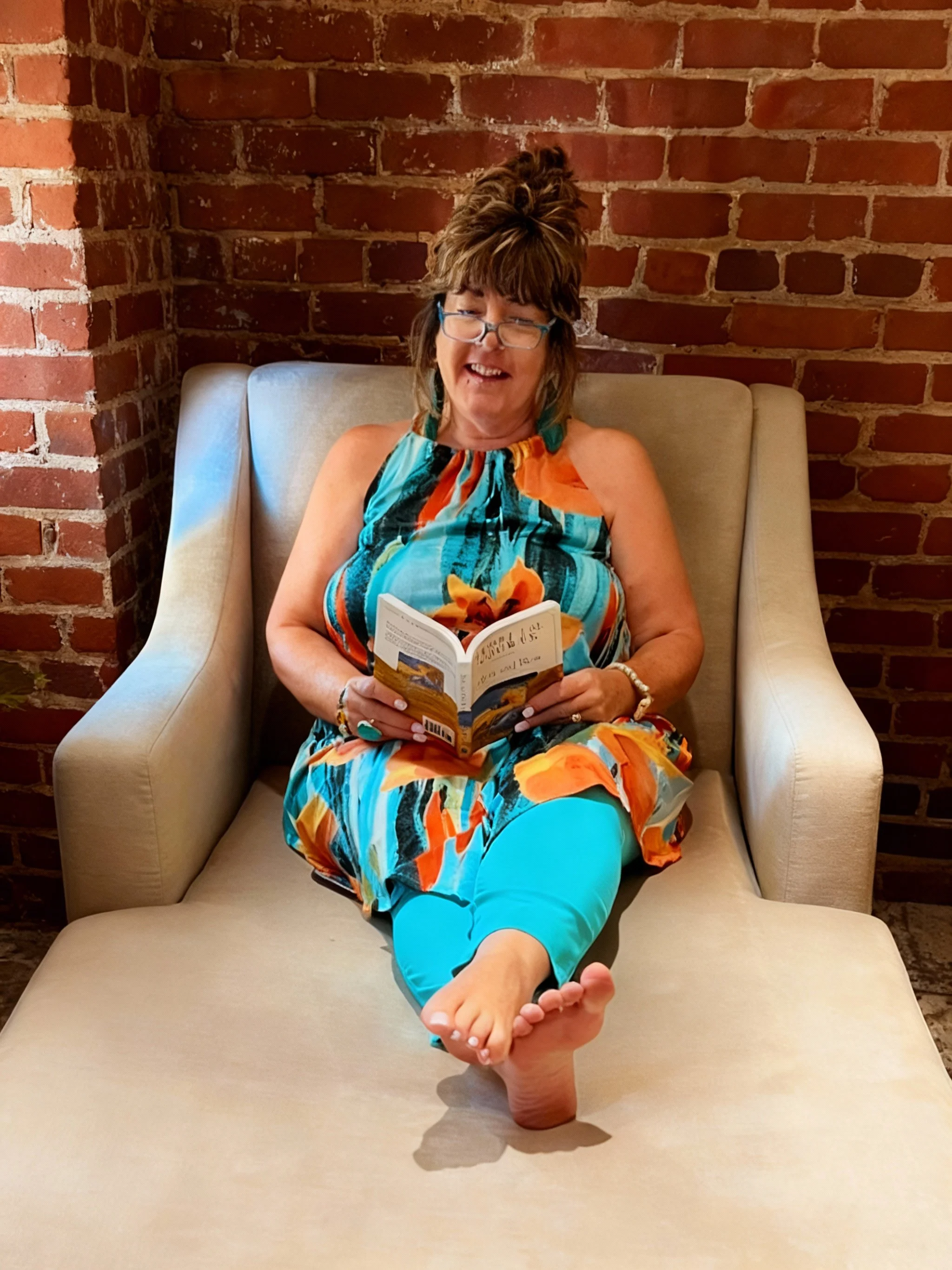 Woman sitting on a beige armchair against a brick wall, smiling while reading a book, dressed in a colorful floral dress and turquoise pants, with her right foot resting on the armchair.