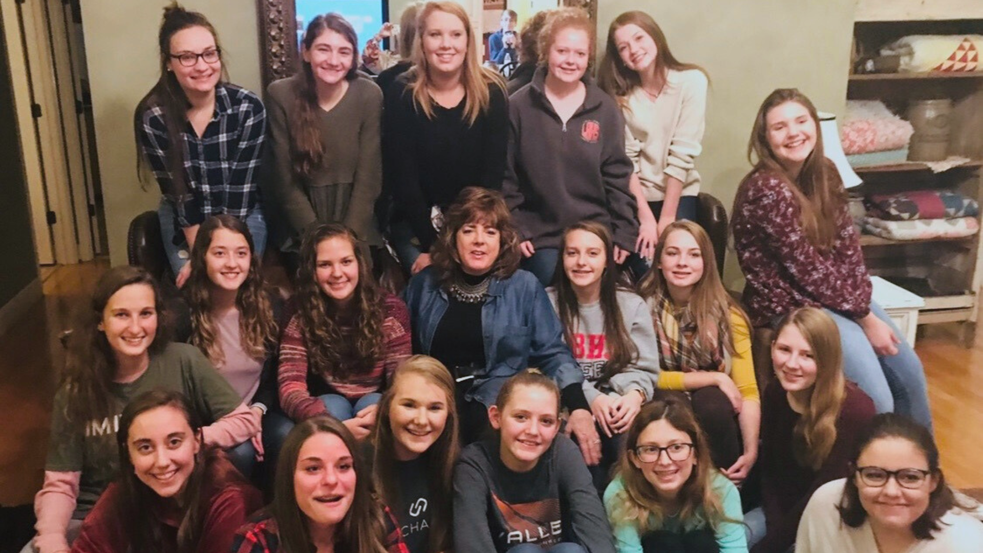 A group of 17 young girls and a woman gathered together indoors for a photo. The girls are smiling and sitting or standing around the woman, who is seated in the center wearing a denim jacket.