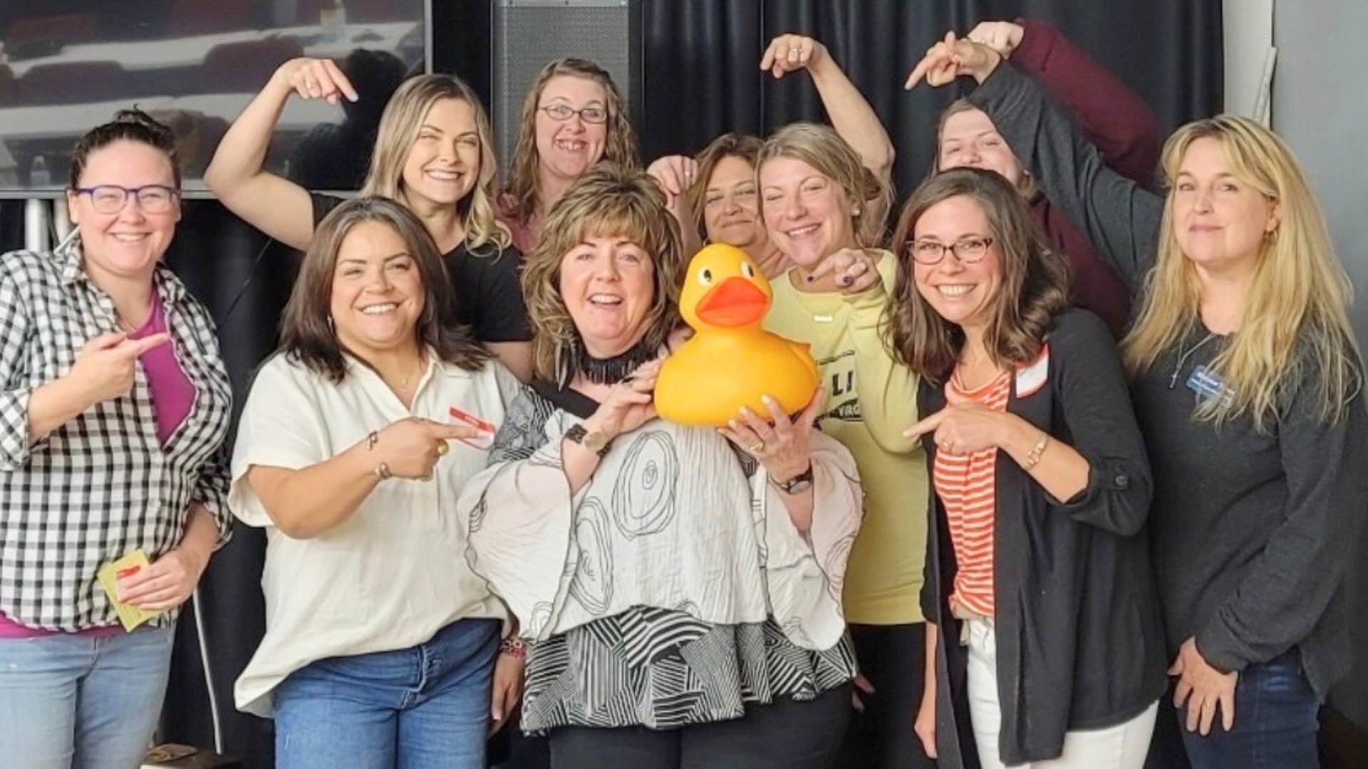 Group of women celebrating, holding a large yellow rubber duck, some pointing and flexing their arms, smiling at the camera.