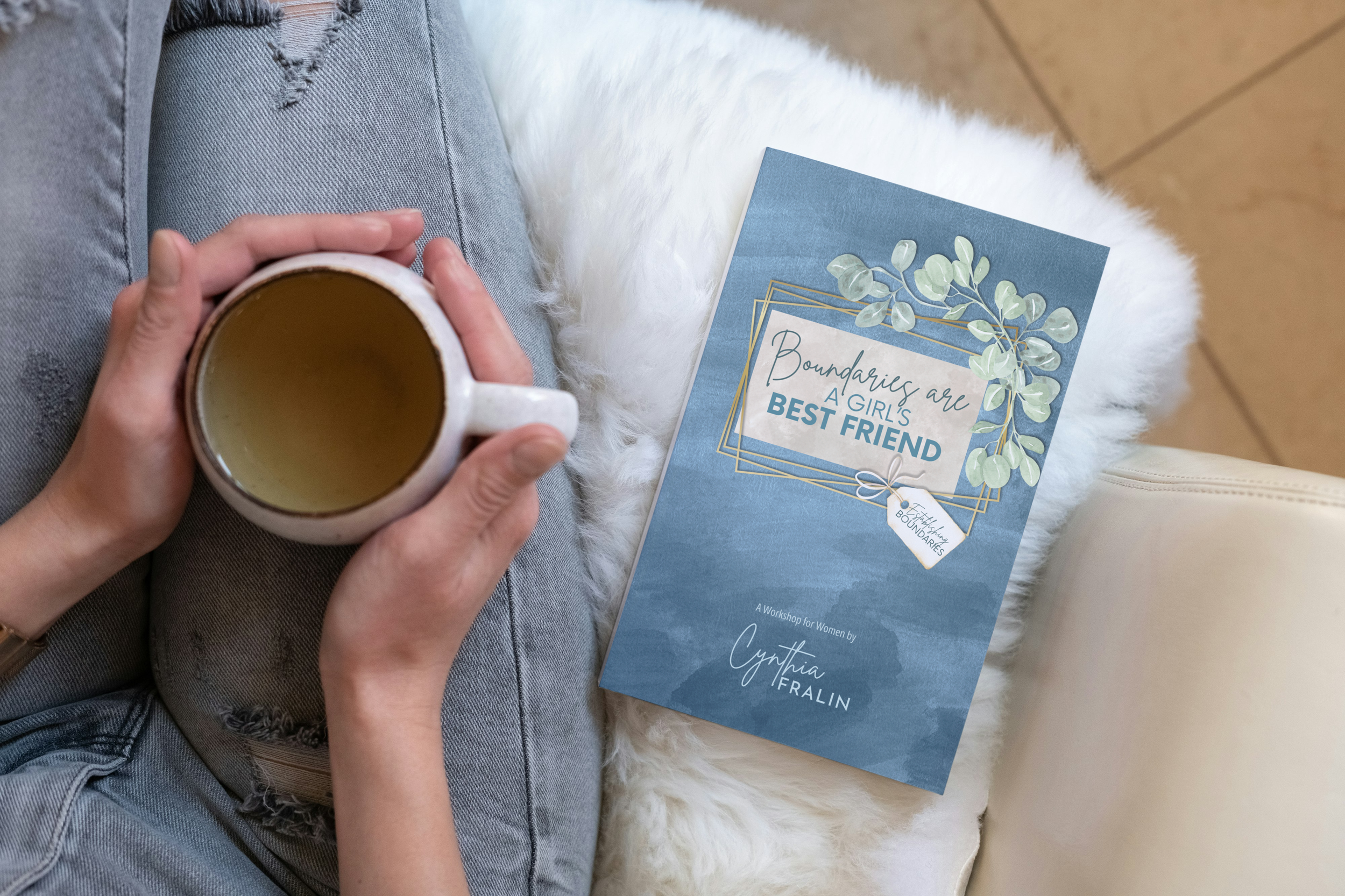 Person holding a mug of tea or coffee, sitting on a soft, white furry blanket with a blue themed booklet titled 'Boundaries are a Girl's Best Friend' by Cynthia Fralin placed beside them.