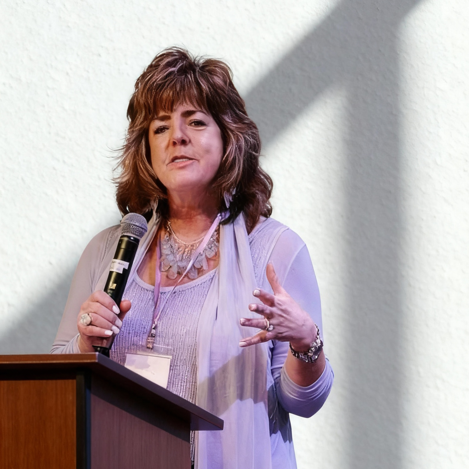 A middle-aged woman with shoulder-length brown hair speaking into a microphone at a podium, wearing a light-colored necklace, bracelet, and a light purple top with a scarf.
