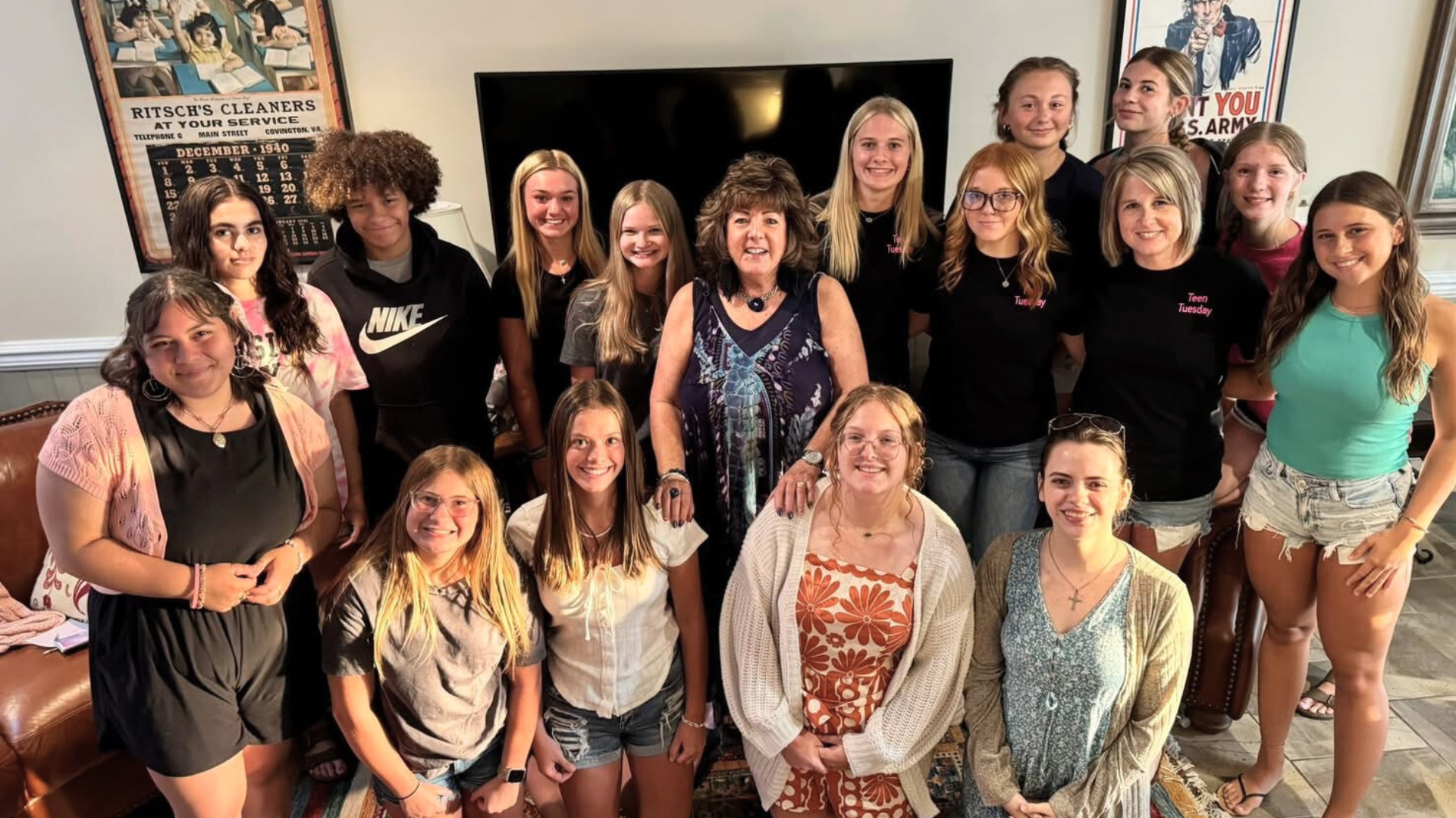 Group of women and girls in a living room, some wearing black shirts with 'Teen Tuesday' embroidered, posing for a photo with a woman in the center.
