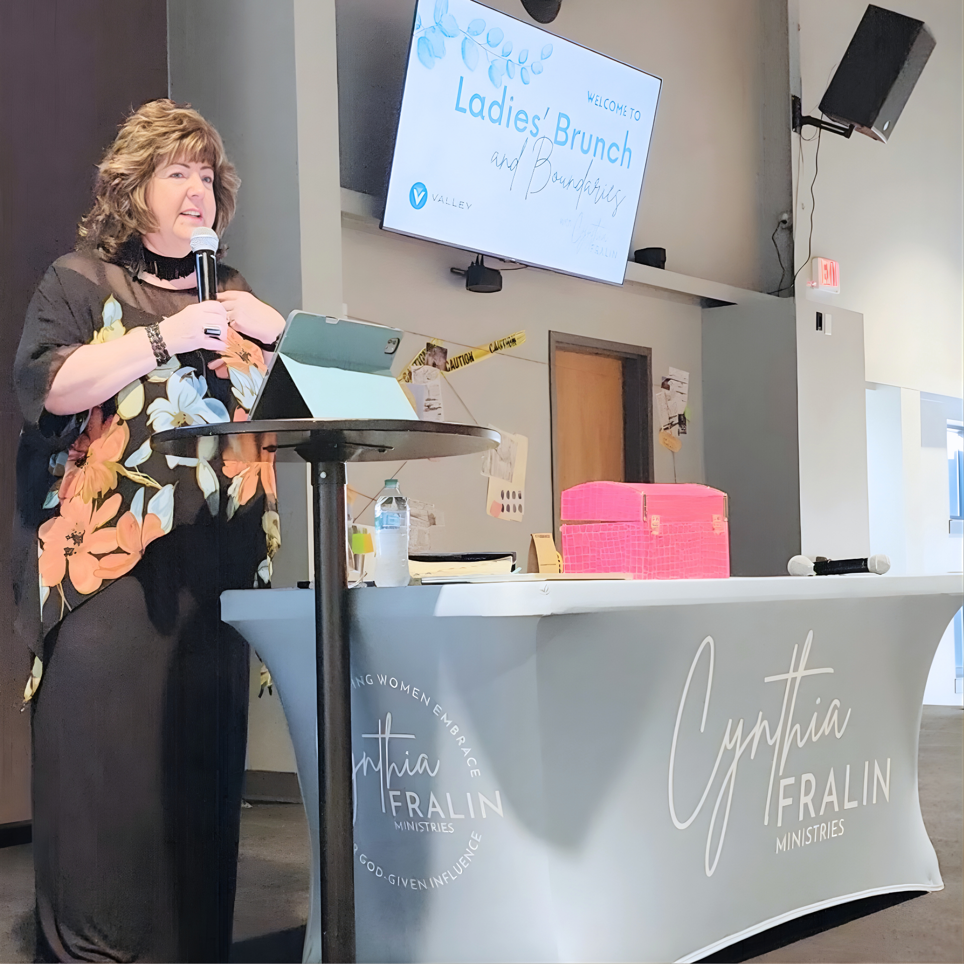 A woman is speaking at a Ladies' Brunch and Boundary event, standing behind a table with a Cynthia Feralin Ministries cloth, items including a water bottle, pink box, and microphone. A screen behind her displays the event details.