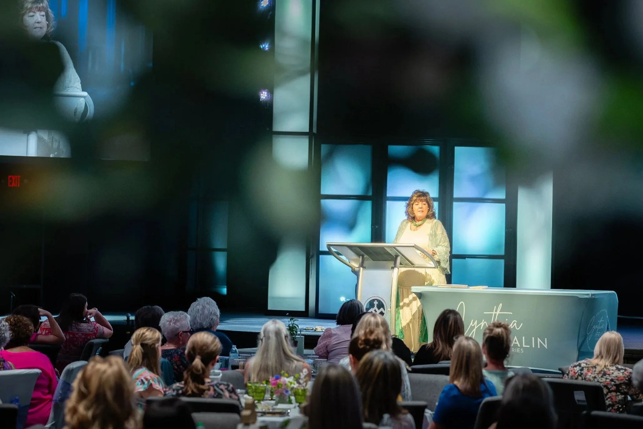 A woman speaking on stage at a conference, with an audience seated at tables in front of her. The stage has a large window backdrop and a decorated table with a sign that reads 'Gytha Kalin.' The audience is diverse, and the woman appears to be giving a presentation or lecture.