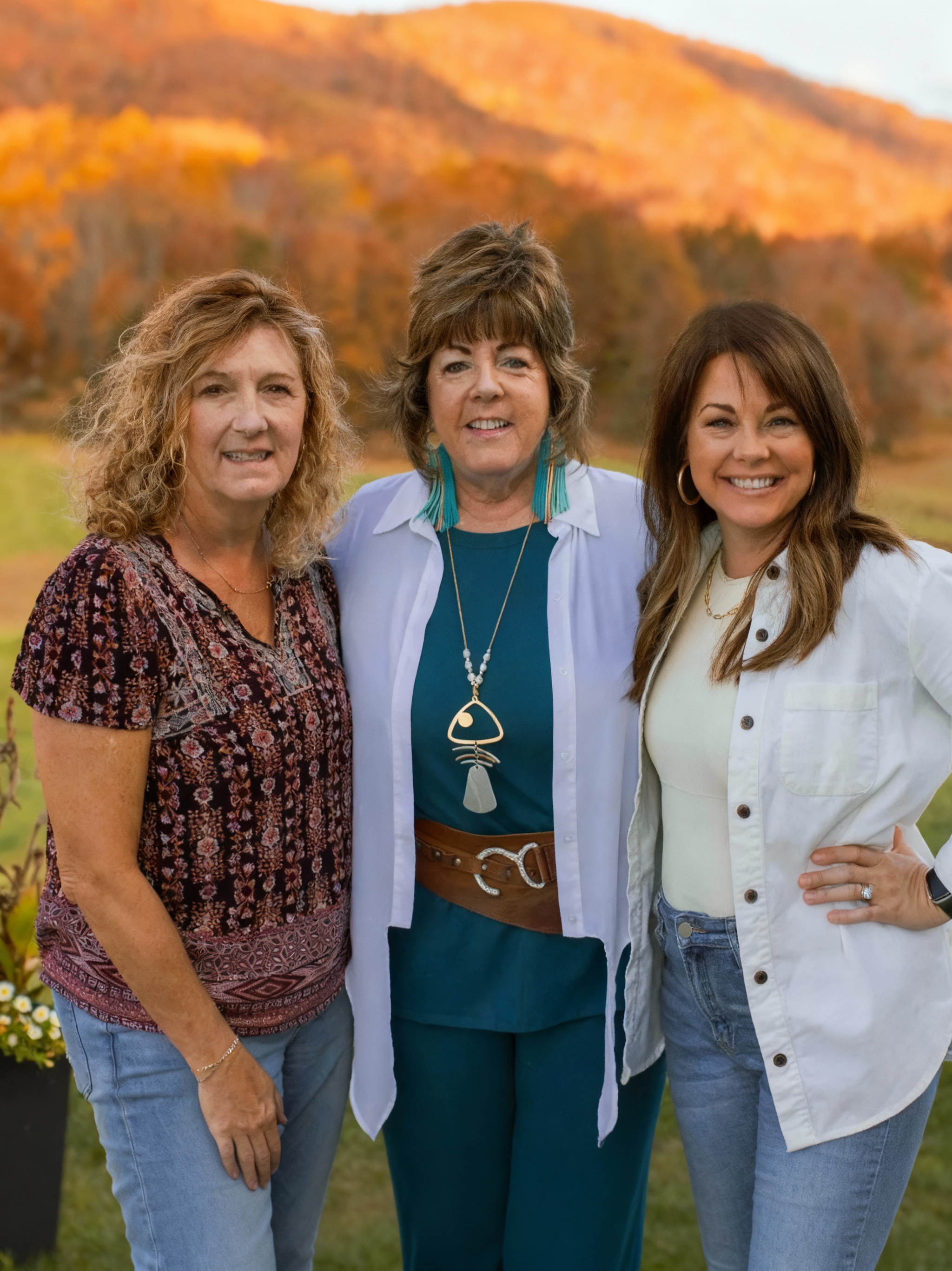 Three women standing outdoors in front of autumn-colored trees, smiling at the camera.