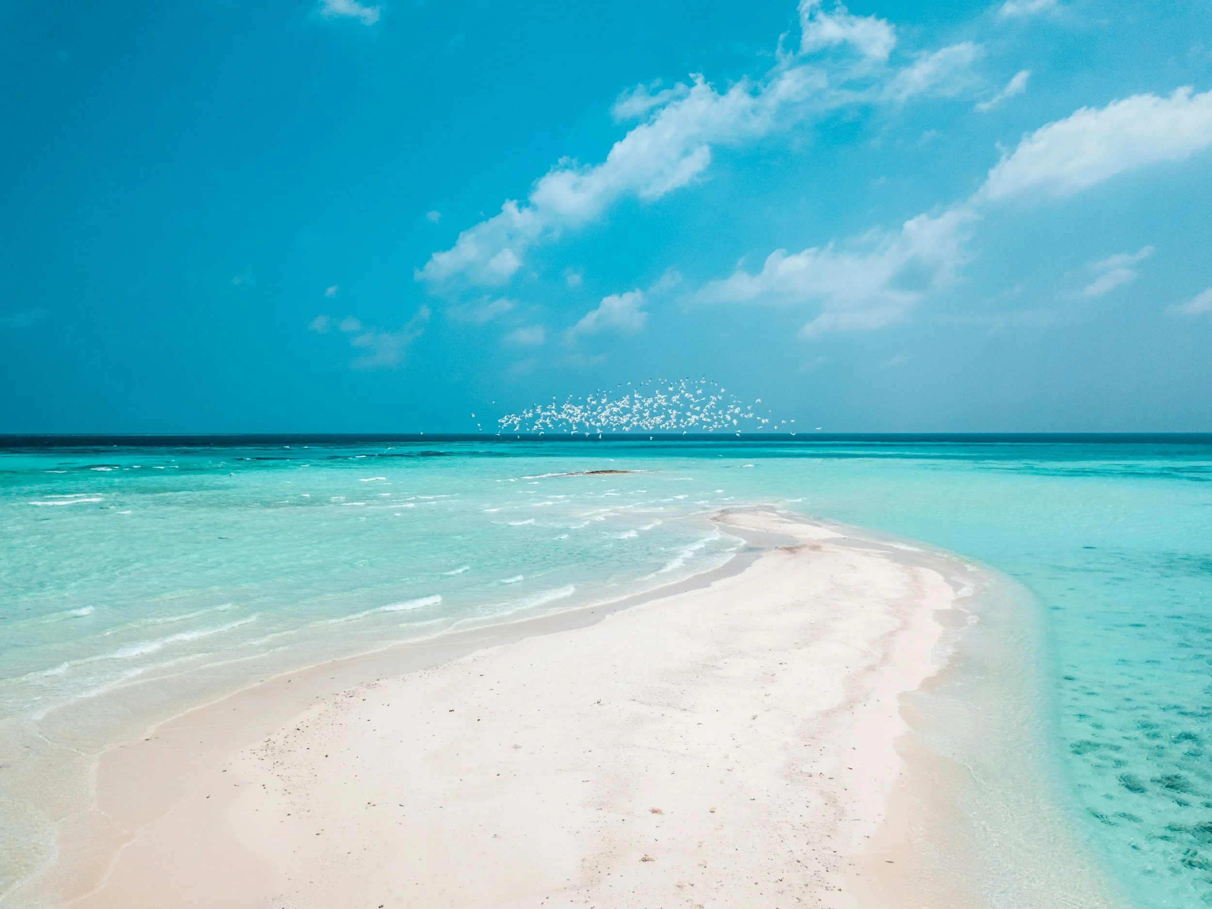 Tropical beach with clear turquoise water, white sandy shore, large rocks, and a blue sky with scattered clouds.