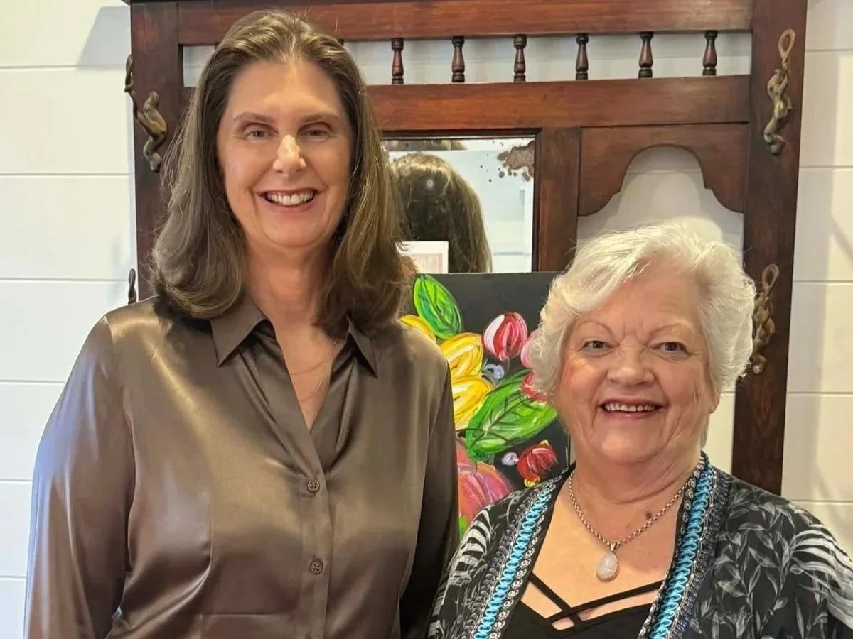 Two women smiling indoors, standing in front of a mirror with a wooden frame, with a colorful painting of flowers behind them.
