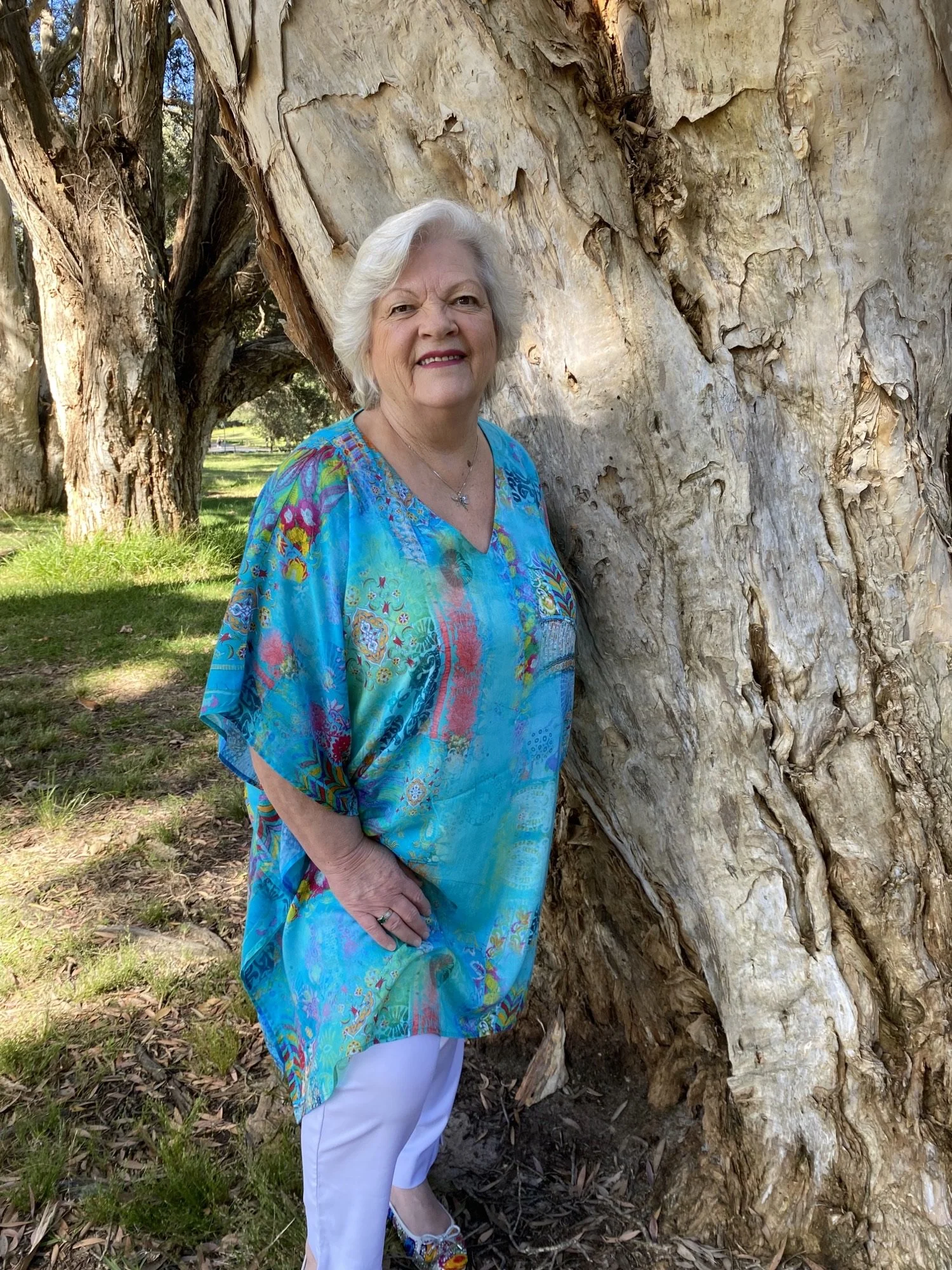 An elderly woman with white hair standing outdoors next to a large, textured tree trunk. She is wearing a colorful turquoise blouse with floral patterns, white pants, and multicolored shoes. The background shows green grass, additional trees, and a clear blue sky.