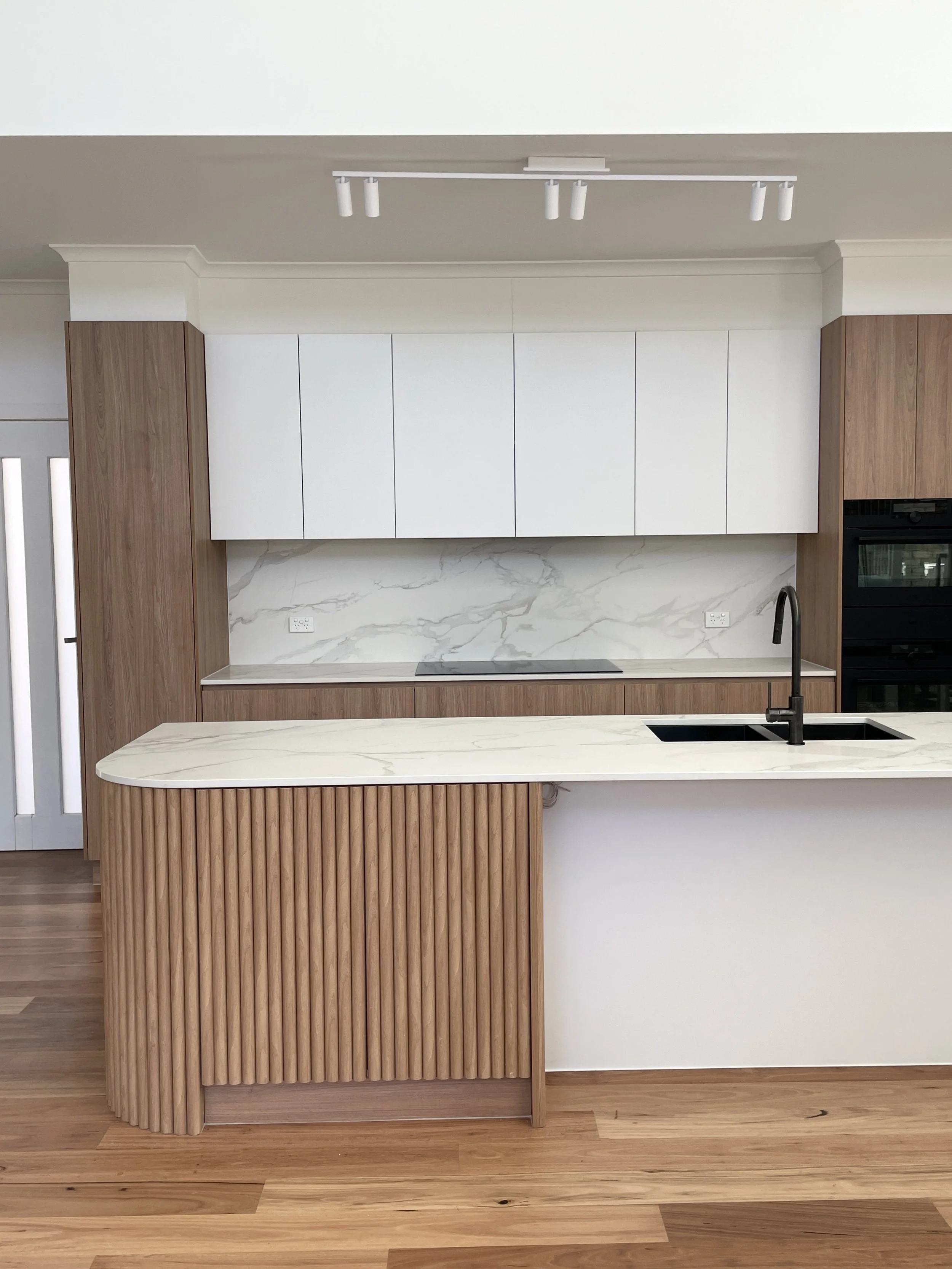 A modern kitchen with white upper cabinets, a marble backsplash, and a wooden island with vertical slats. There is a black faucet over a double sink and a black oven on the right side.