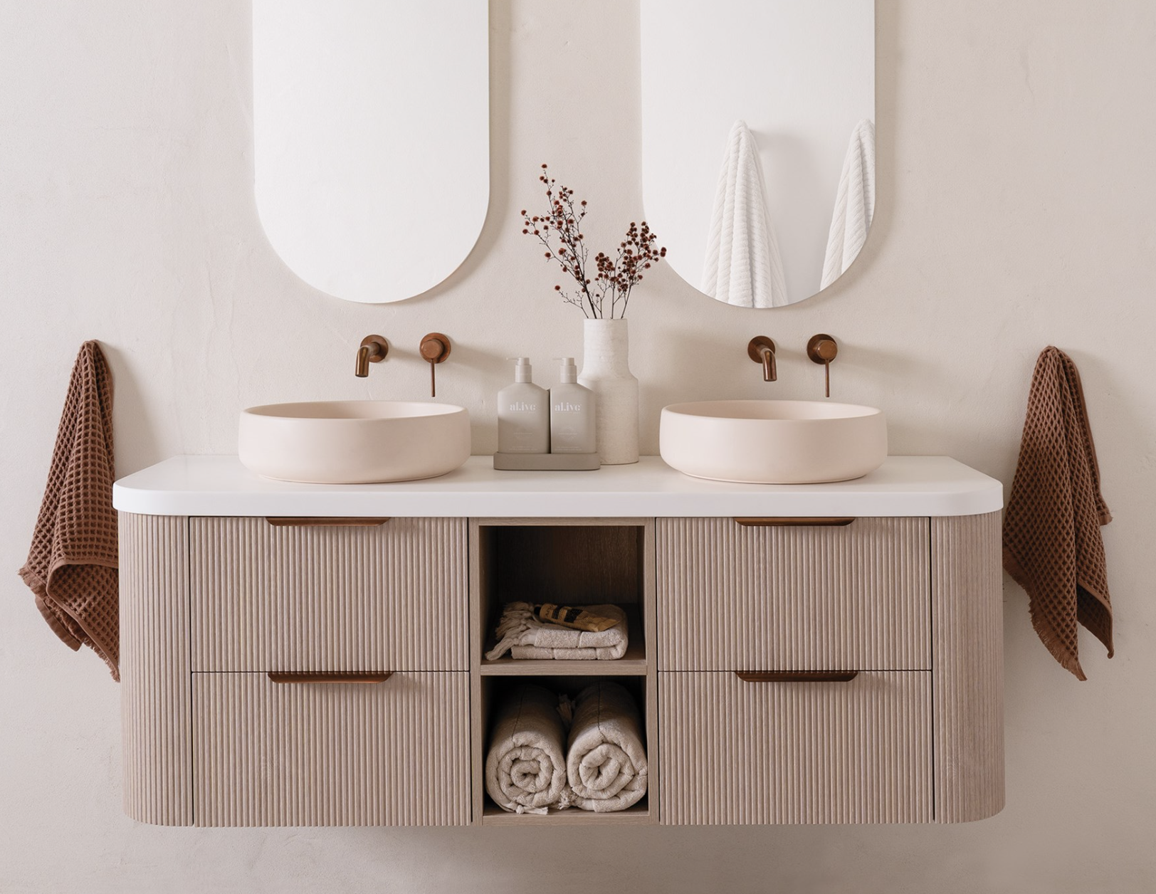A modern bathroom vanity with two round vessel sinks, two mirrors, and decorative elements like towels, soap dispensers, and a vase with branches.