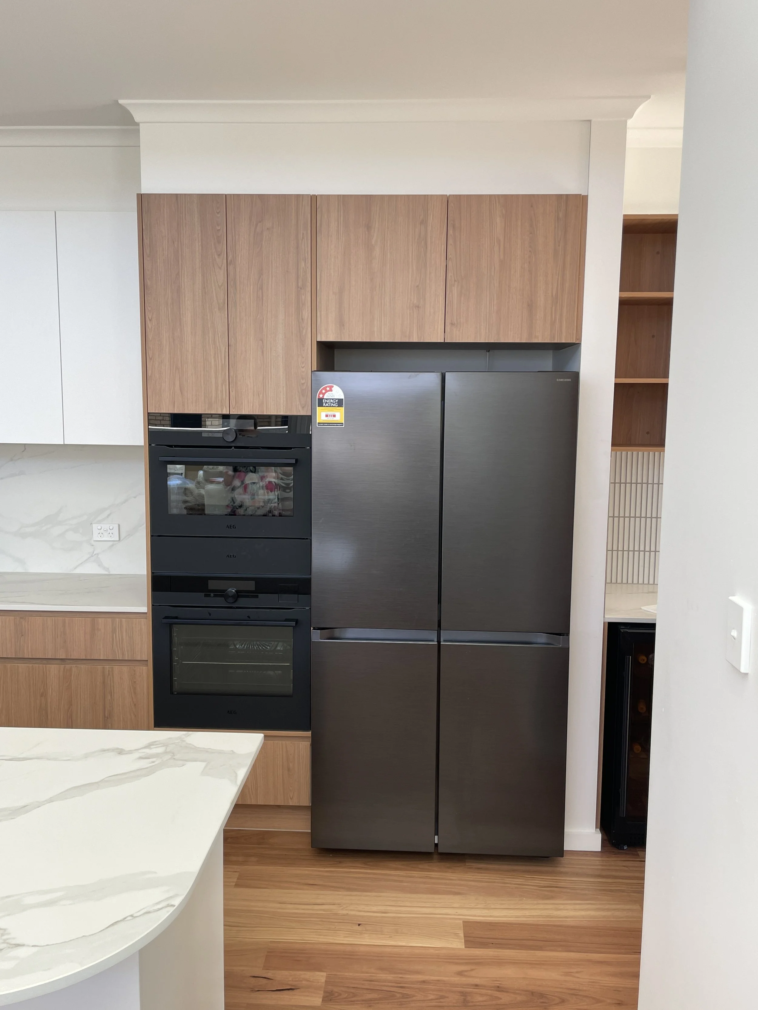 Modern kitchen with white marble countertop, black oven, and stainless steel refrigerator, beige wooden cabinets, and a built-in wine rack.