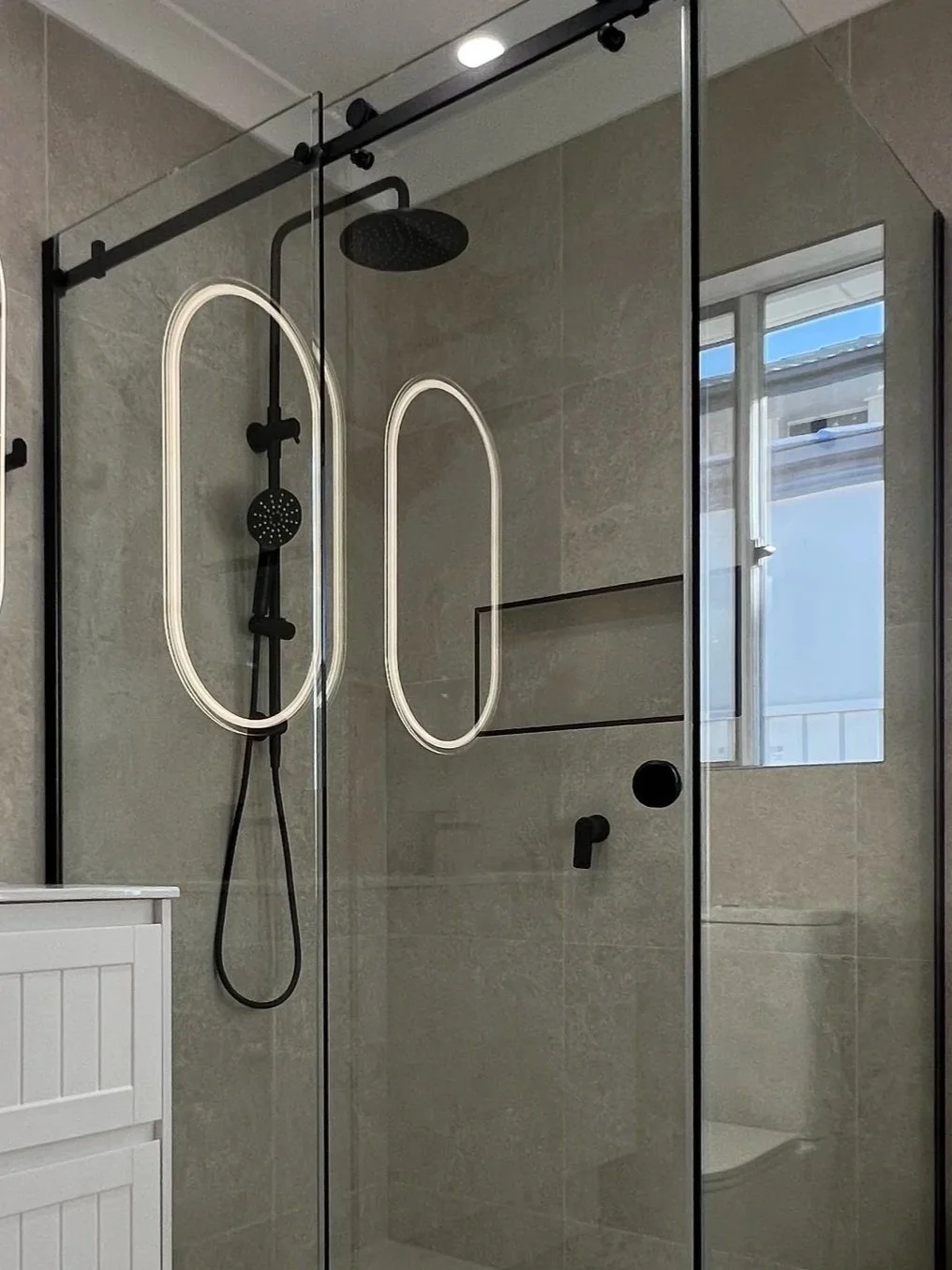 Modern glass shower with black fixtures and a window showing a building outside.
