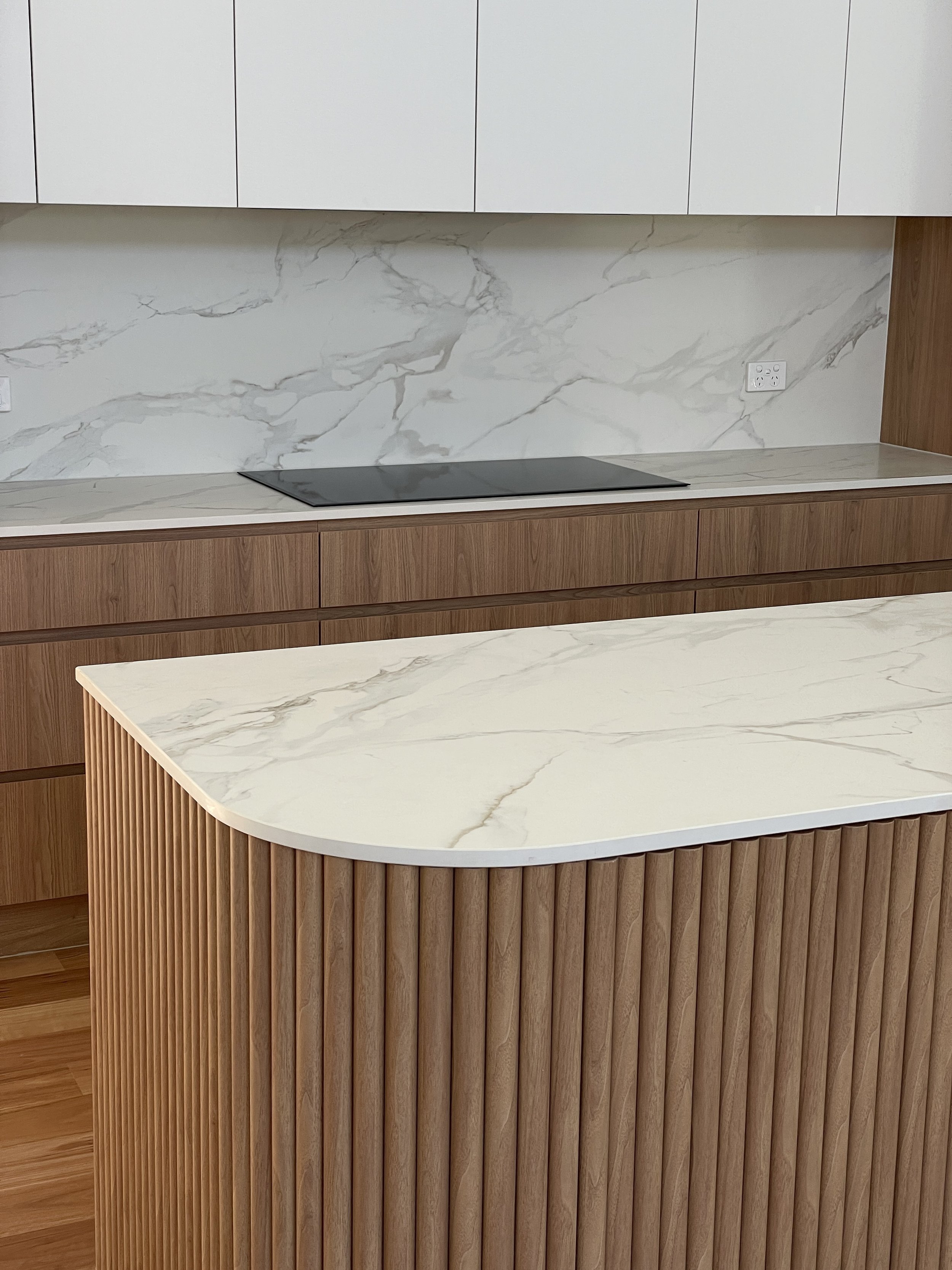 Modern kitchen with a white marble countertop, wooden lower cabinets, white upper cabinets, and a black induction stovetop, with electrical outlets on the wall.
