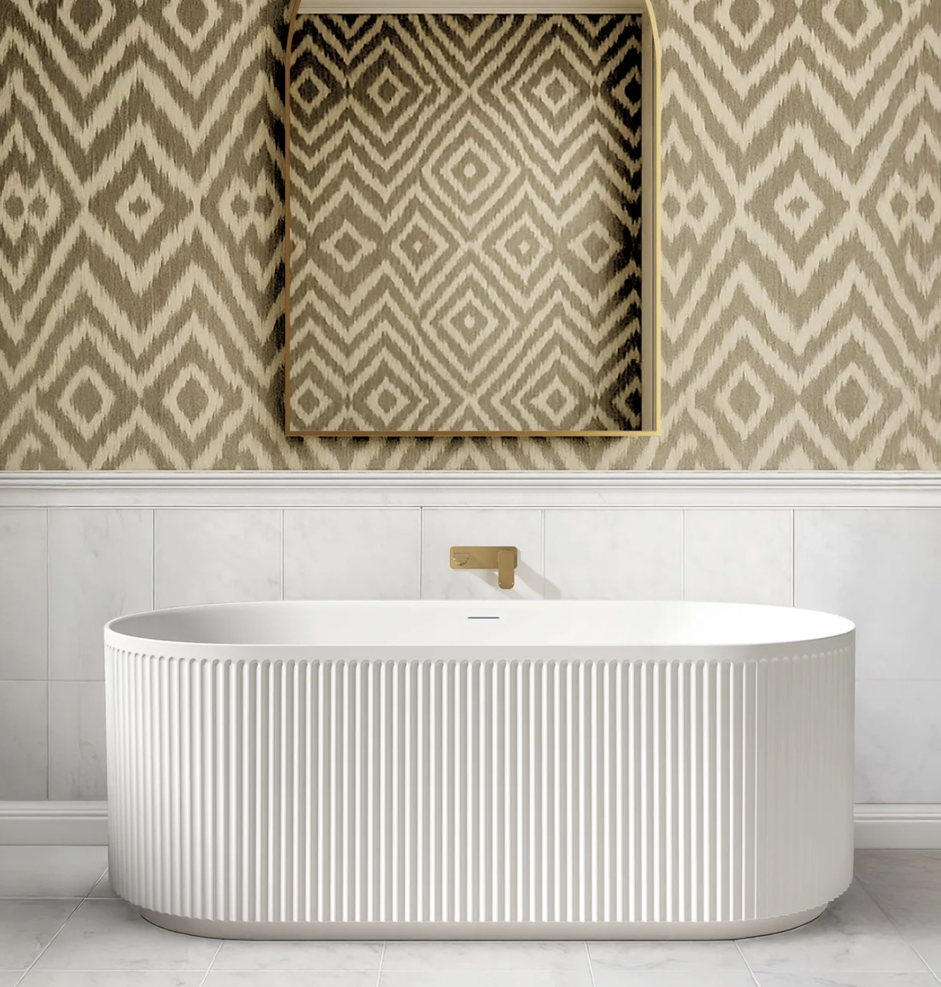 Modern bathroom with a freestanding white bathtub with vertical ridges, a gold faucet, a large mirror with a gold frame on a patterned beige wallpaper wall, and white tiled lower wall with a white tile floor.