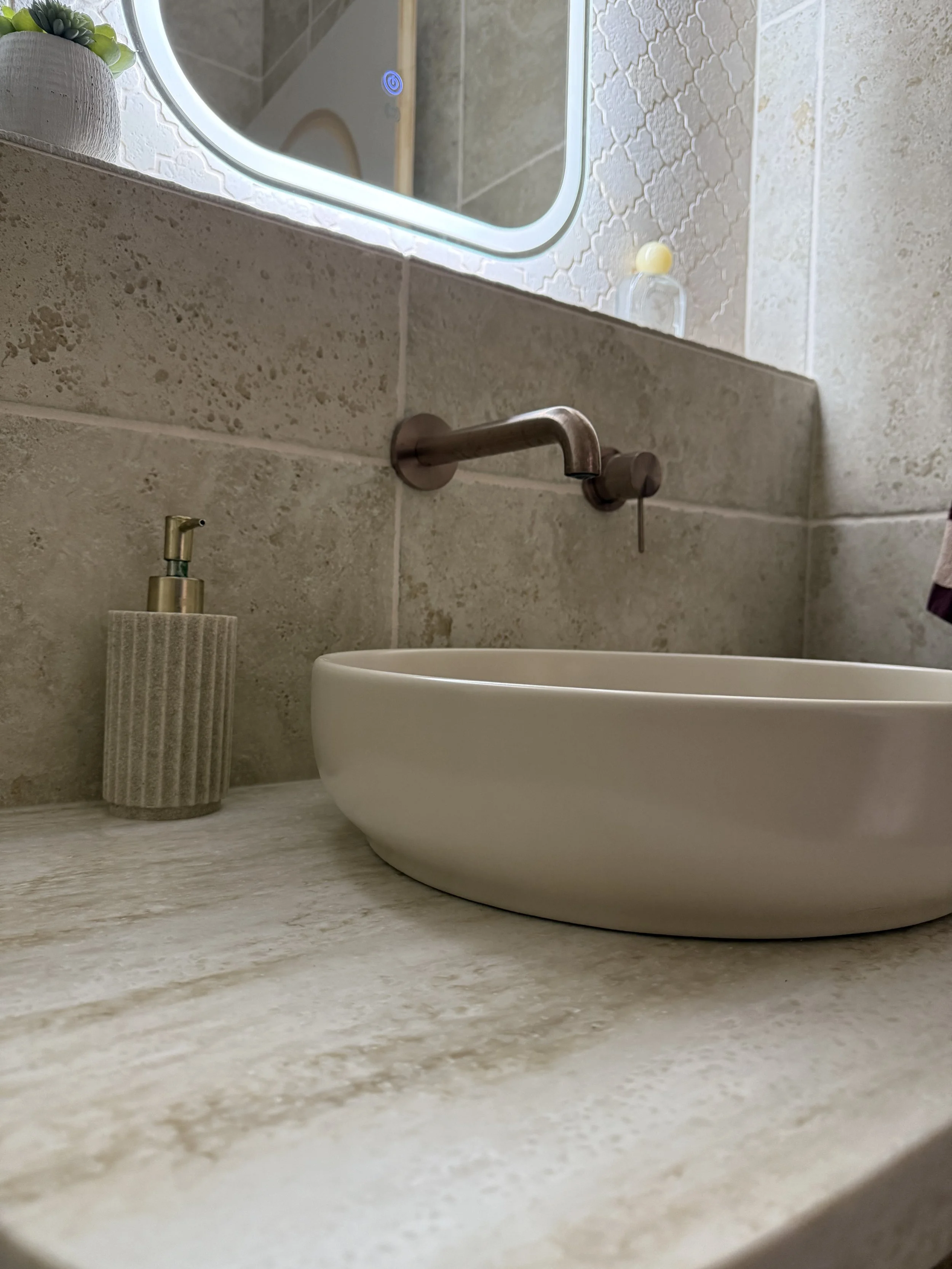 A bathroom countertop with a white ceramic sink, a wall-mounted faucet, a soap dispenser, a mirror with a built-in light frame, and part of a soap bottle visible. The wall behind has textured tiles and a small plant in a pot.