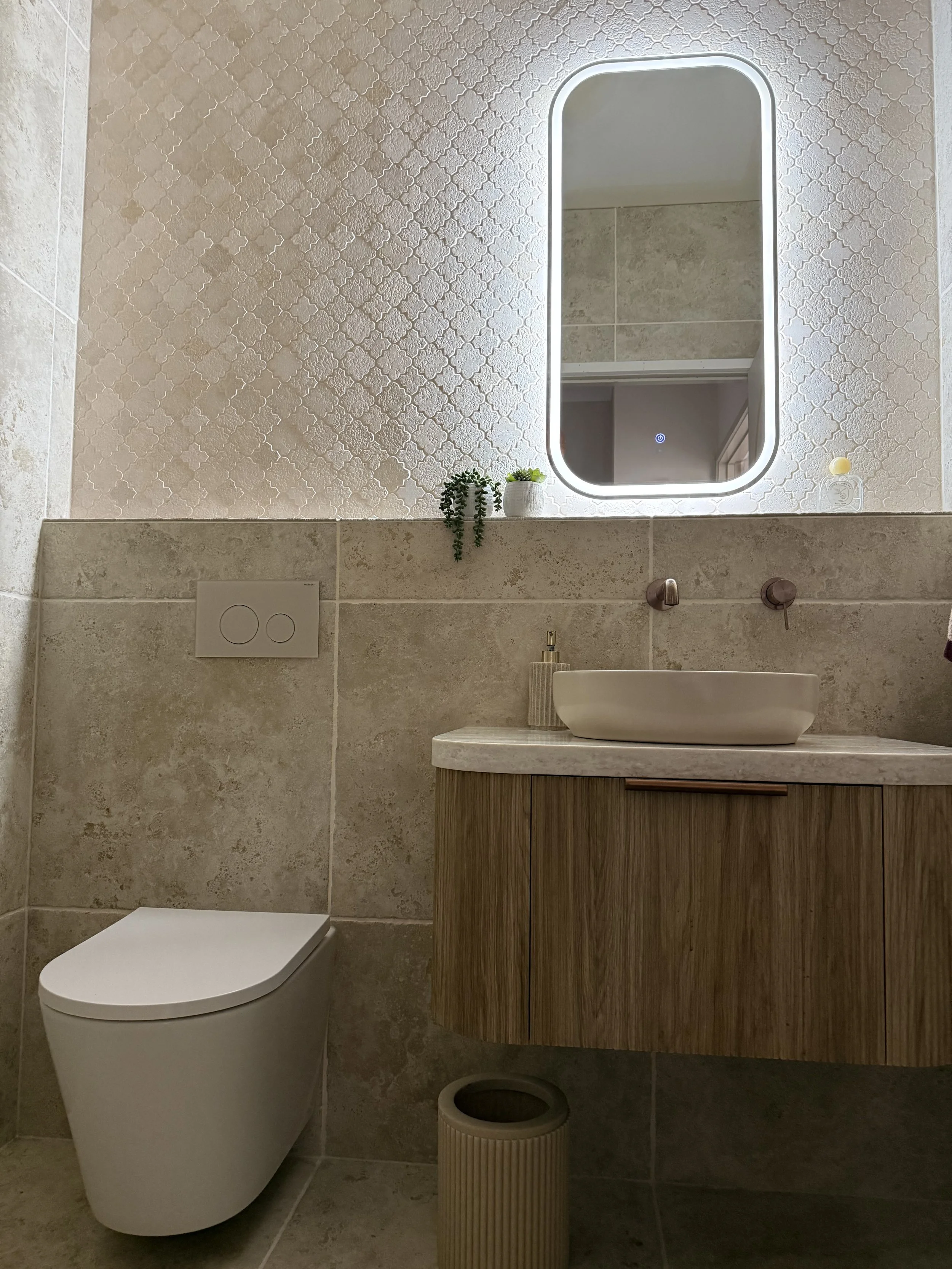 Contemporary bathroom featuring a wall-mounted toilet, a wooden vanity with a vessel sink, a mirror with LED lighting, and small decorative plants on the shelf above the sink.