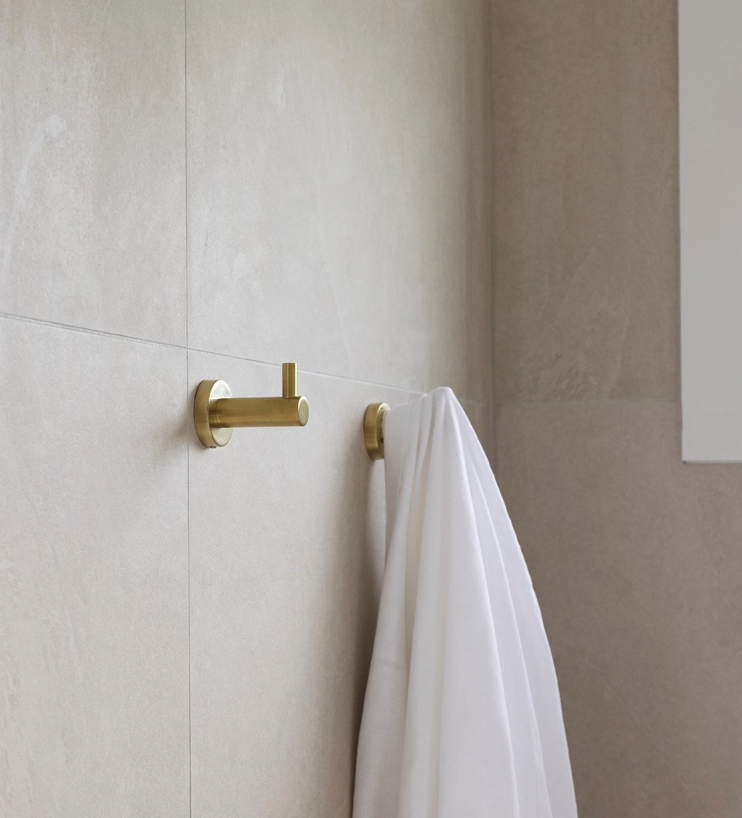 A white towel hanging on a gold towel hook on a beige tiled wall in a bathroom.
