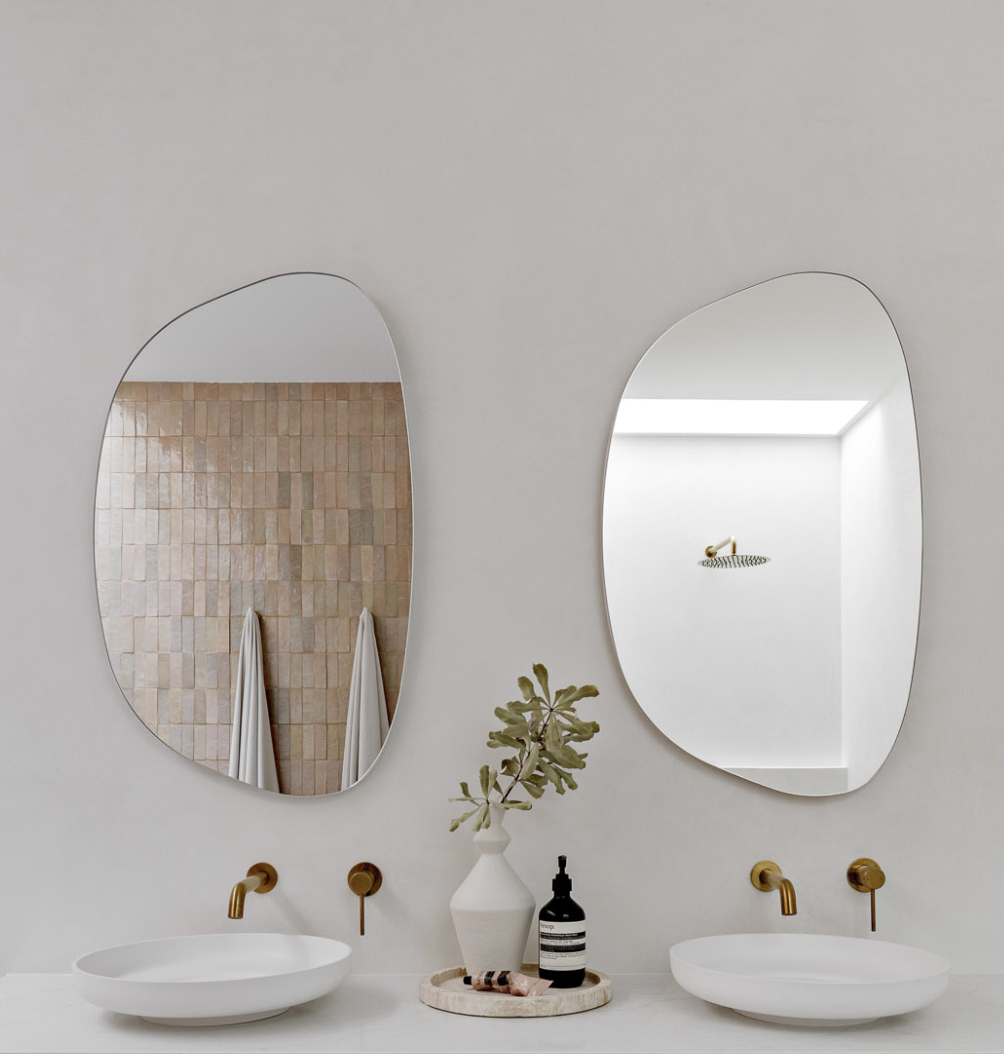 Modern bathroom with two oval mirrors above white sinks, gold fixtures, and minimal decor, including a vase with greenery and a black bottle on a round tray.
