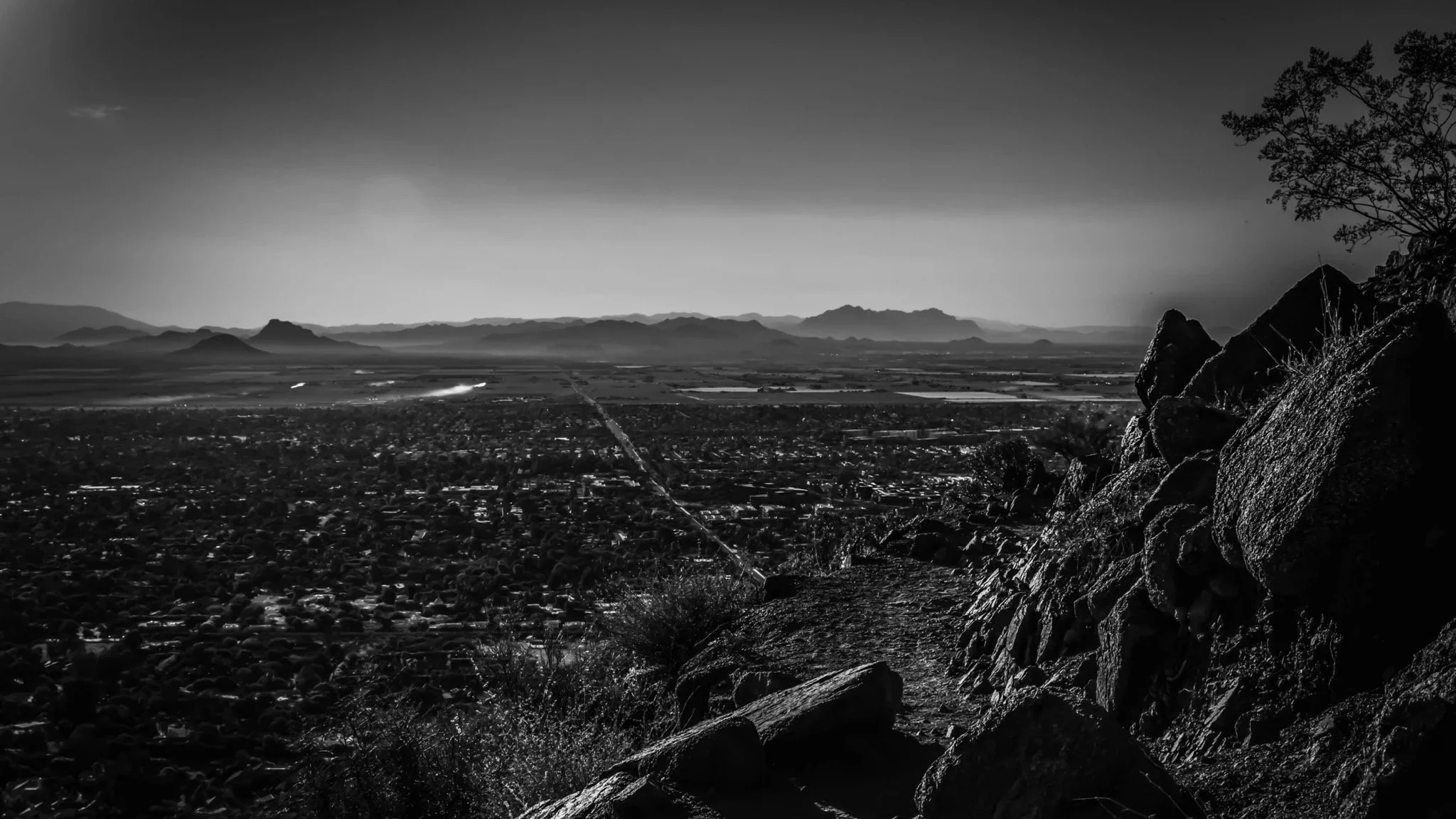 A black and white photograph of a cityscape seen from a mountain trail with rocks and a tree in the foreground, and mountains in the distance.