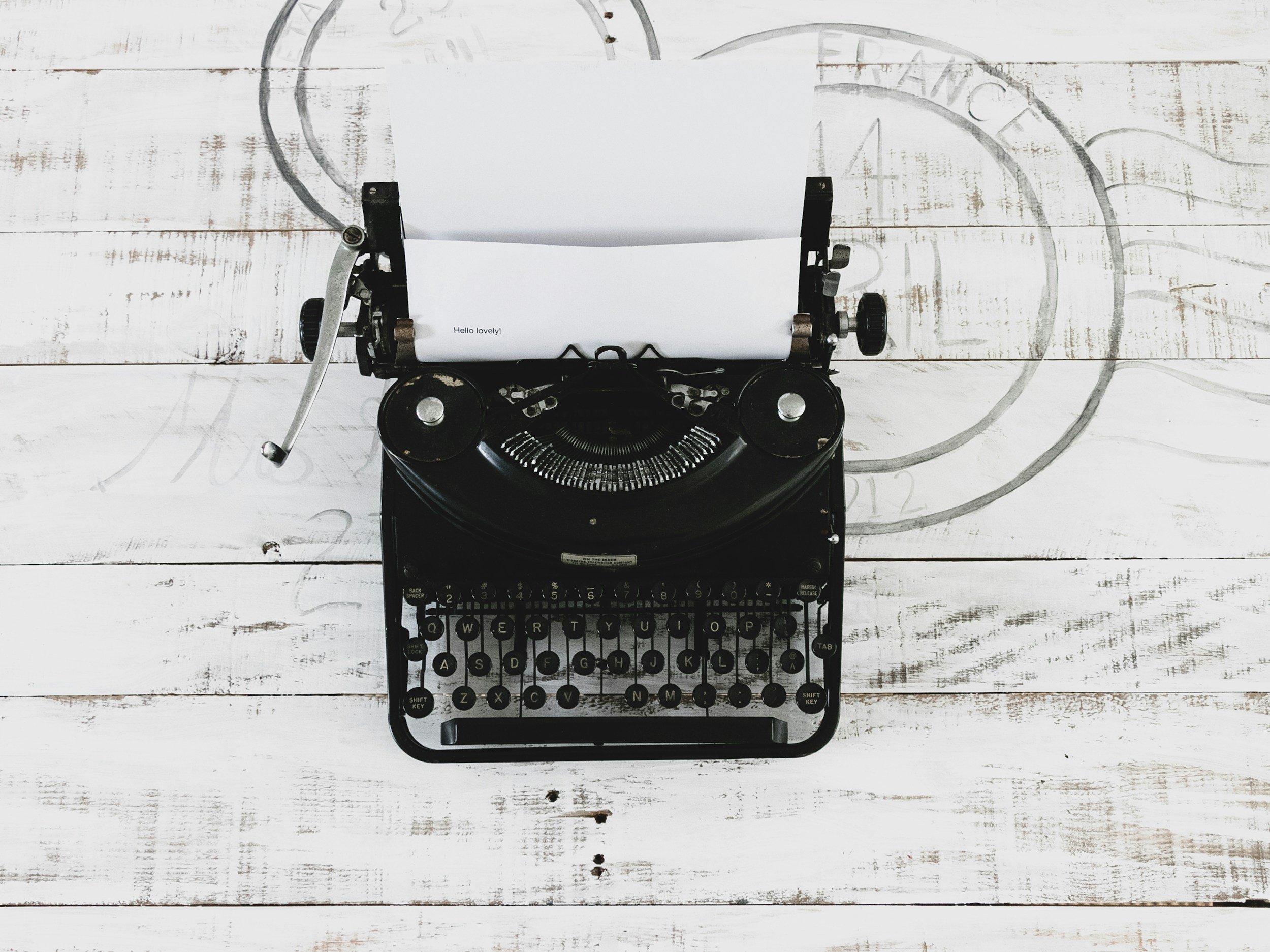 A vintage black typewriter sitting on a white wooden surface, with a sheet of paper in the roller that has the text 'Hello lovely!' on it.