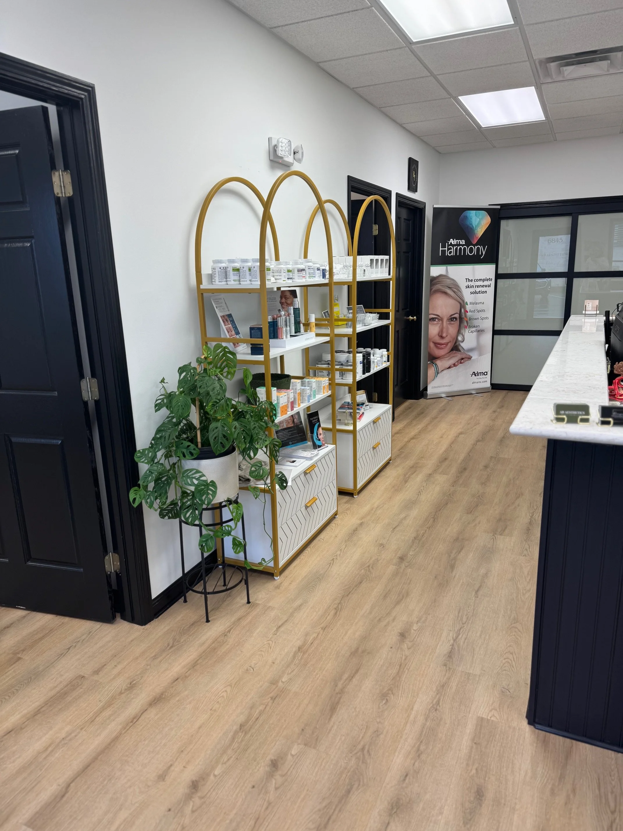 Interior of a modern medical or skincare clinic with white walls, wood flooring, black door frames, and a reception counter. There are two gold shelving units with skincare products and a large green potted plant on a black stand next to the shelves. A promotional stand with a woman's face and the word 'Harmony' is visible in the background.