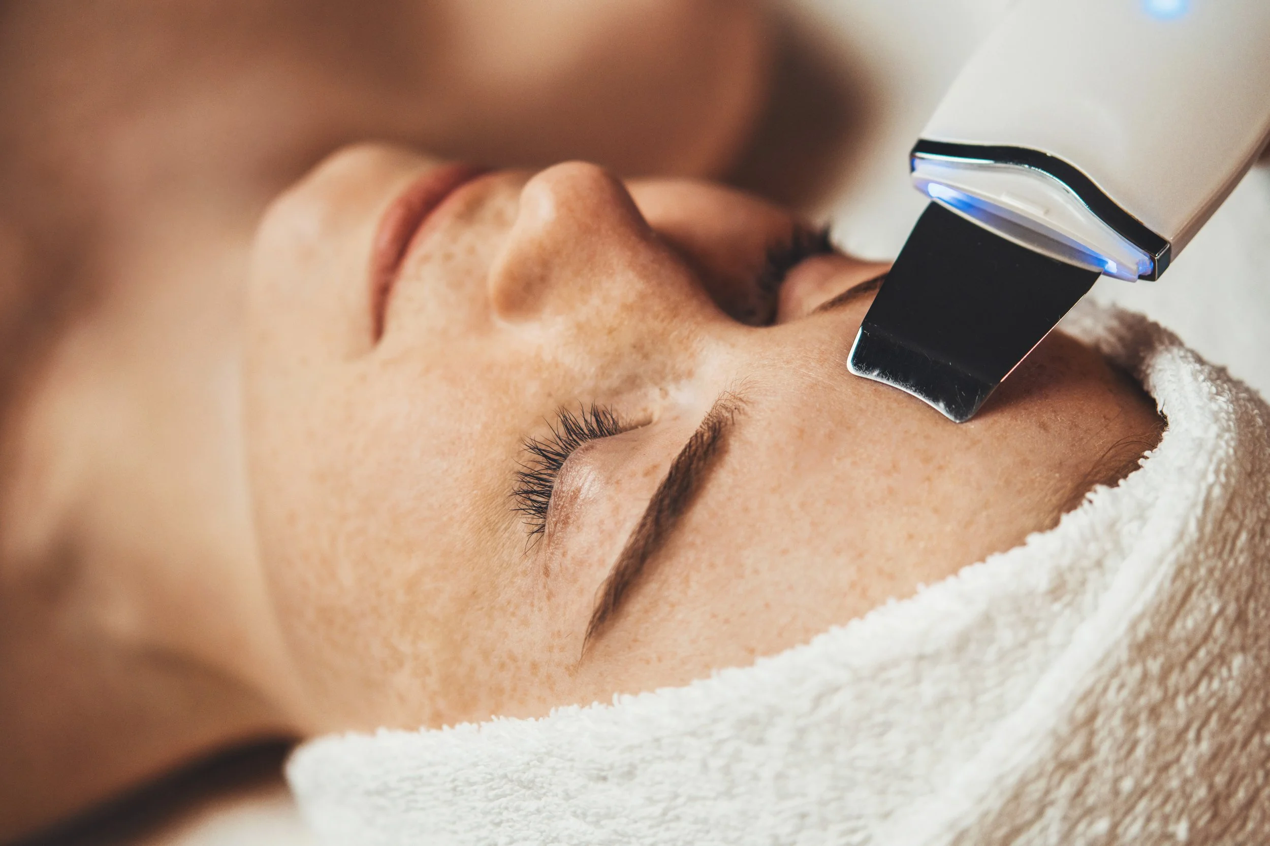 A woman receiving a facial treatment, with her eyes closed, in a spa or skincare clinic, while a device is positioned near her nose.