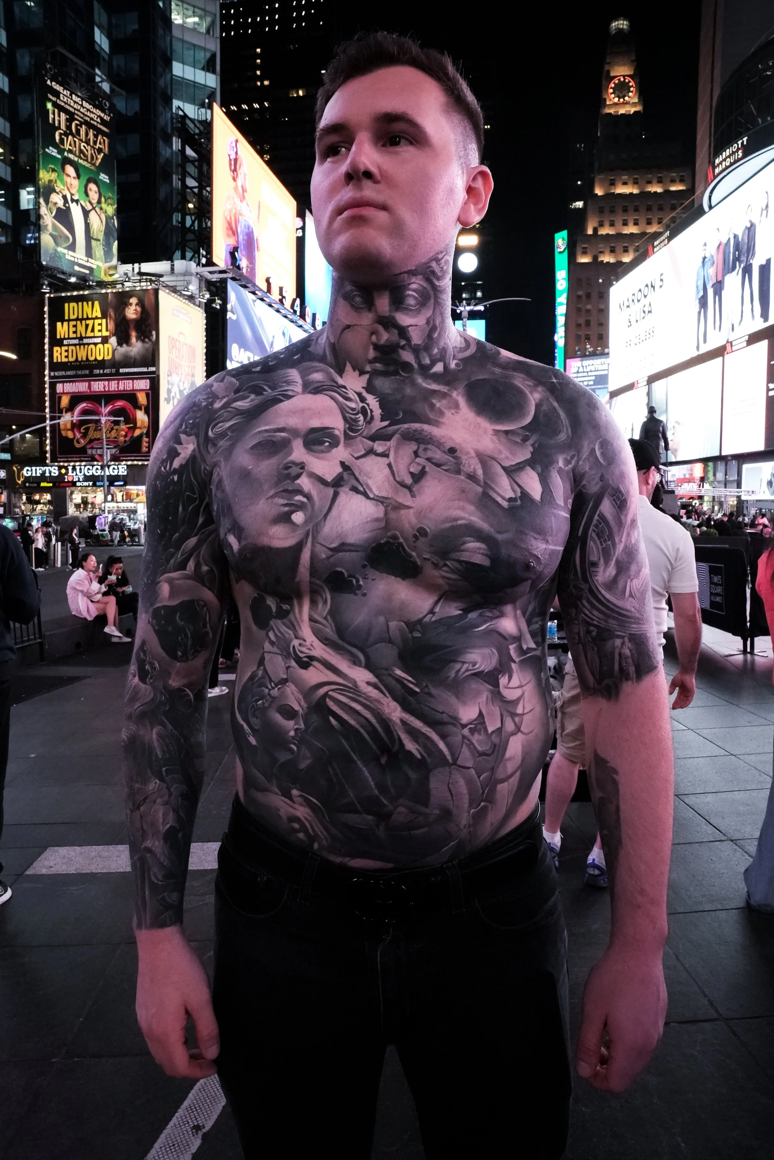 Living room with colorful artwork and patterned ceiling. Features a large gray sofa with geometric and face-patterned throw pillows, a dining table with chairs, plants, large windows, and a black-and-white mural on the ceiling.