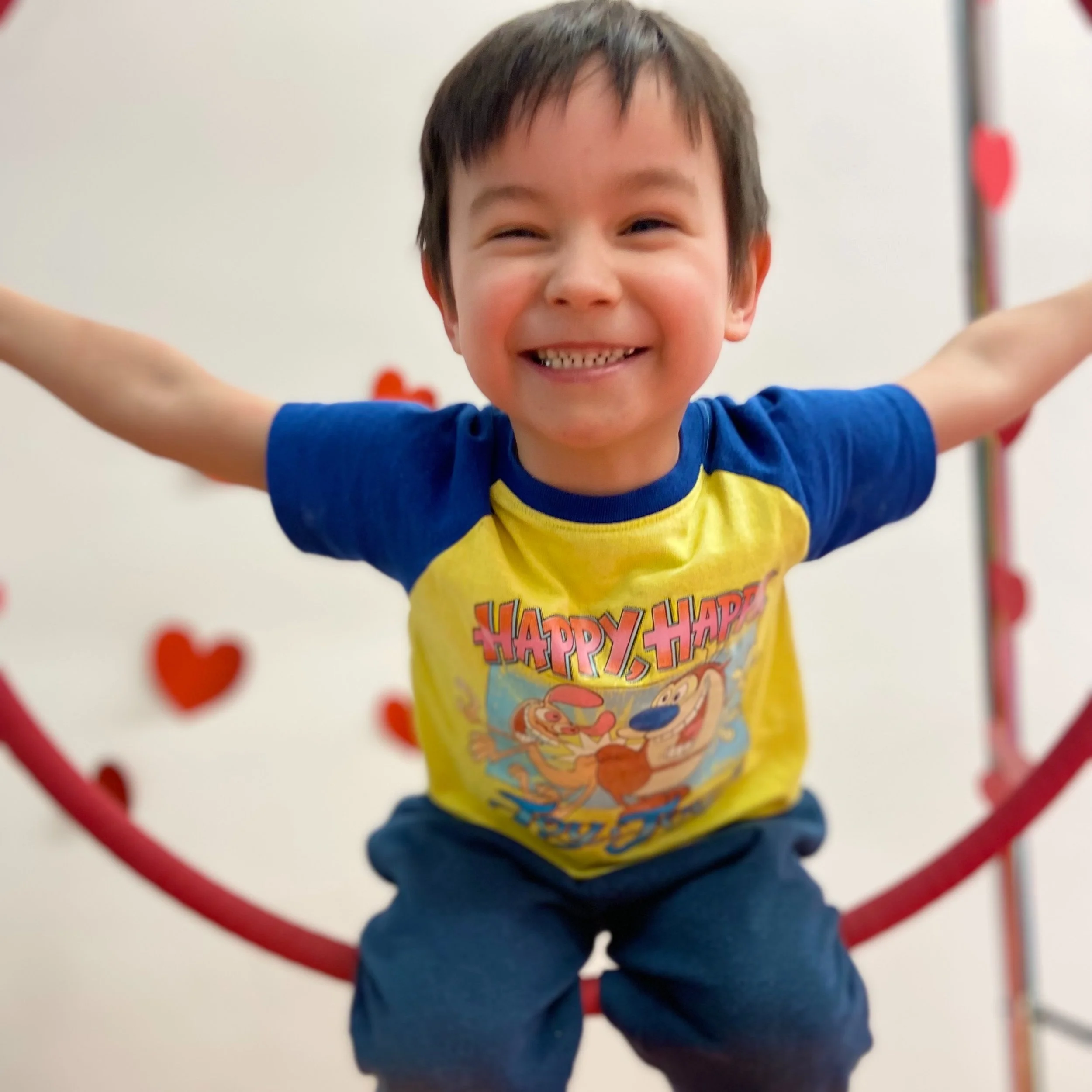 Smiling child in blue and yellow Ren & Stimpy shirt sitting in a red hoop with heart decorations in the background.