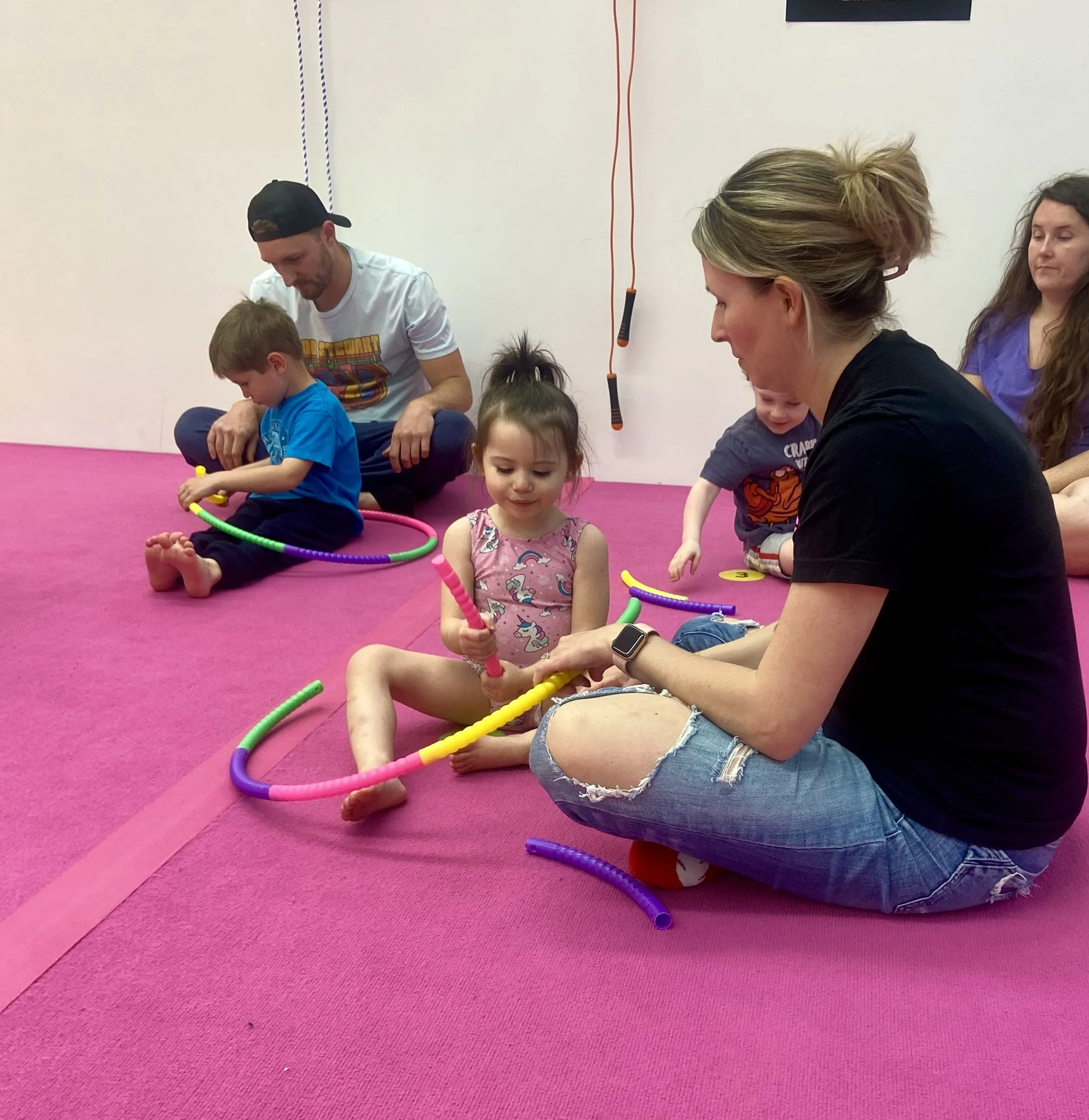Children and adults sitting on a pink mat with colorful foam tubes, engaging in an activity together.