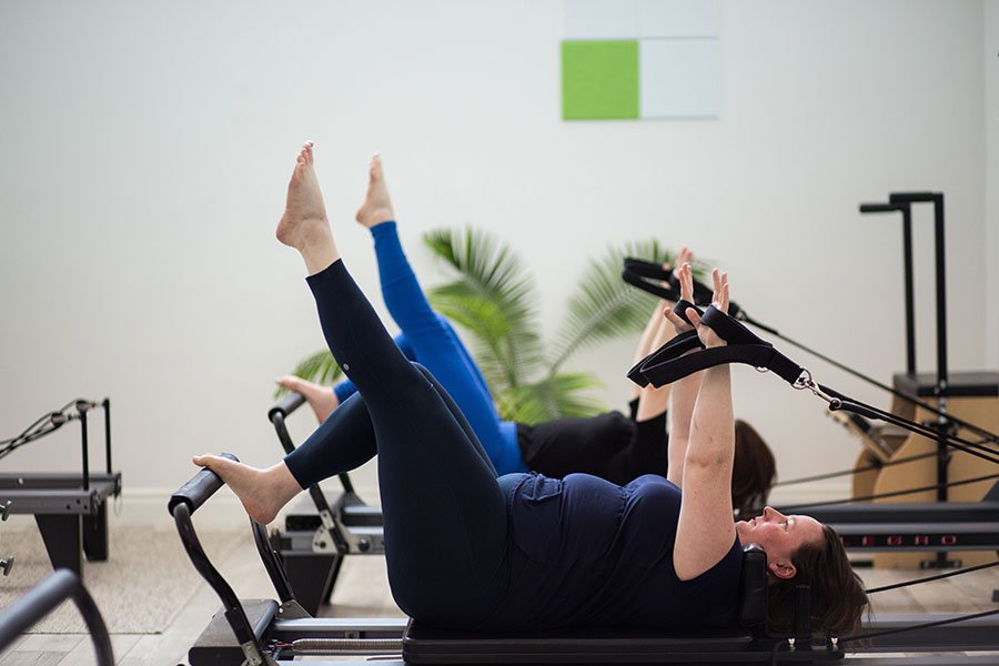 Women participating in a Pilates class using reformer machines with resistance straps, in a sunny fitness studio.
