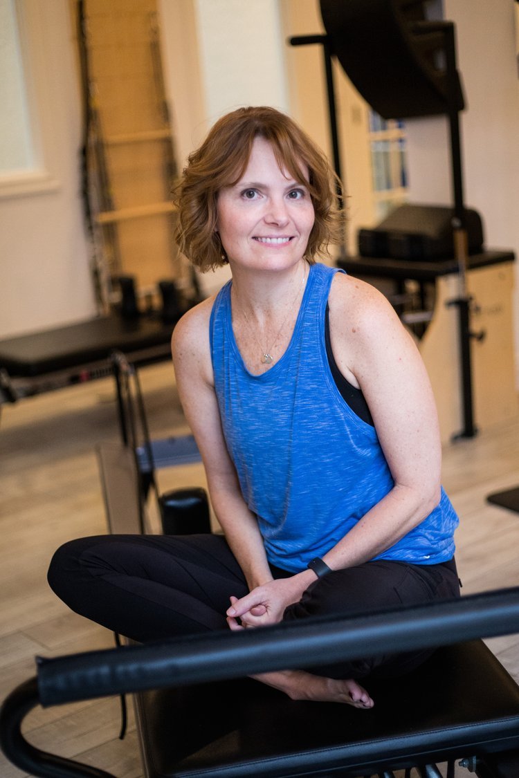 A woman wearing a blue tank top and black pants sitting cross-legged indoors, smiling at the camera.